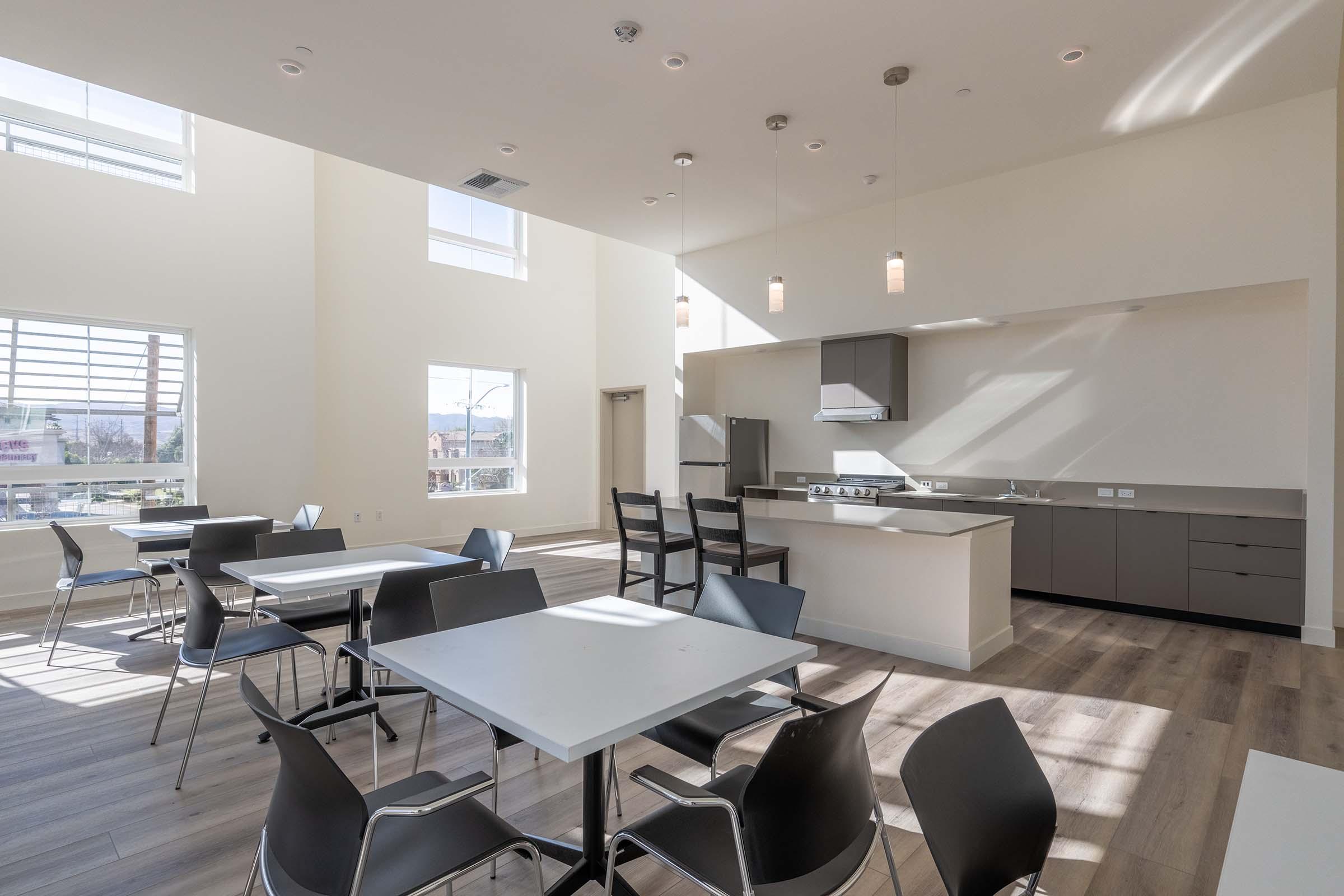 Modern communal dining area featuring several white tables with black chairs. The space has high ceilings, large windows allowing natural light, and a contemporary kitchen area with gray cabinets and appliances. Minimalist design creates an open, airy atmosphere.