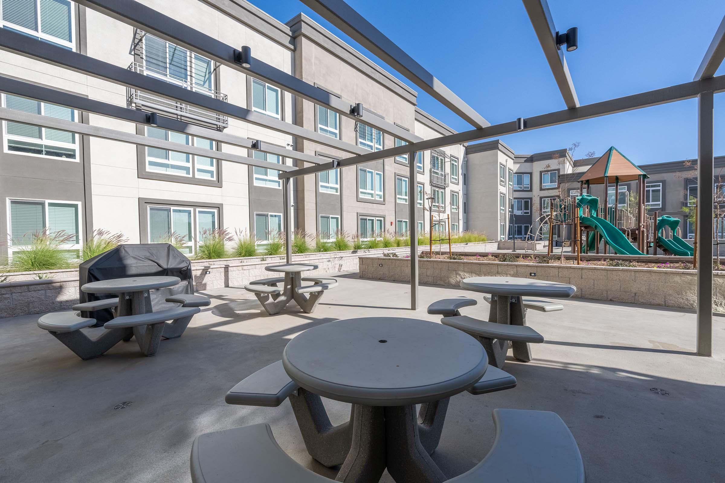 Outdoor seating area featuring multiple picnic tables under a metal pergola. In the background, there is a playground with slides and climbing structures. The setting is surrounded by modern apartment buildings and greenery, all under a clear blue sky.