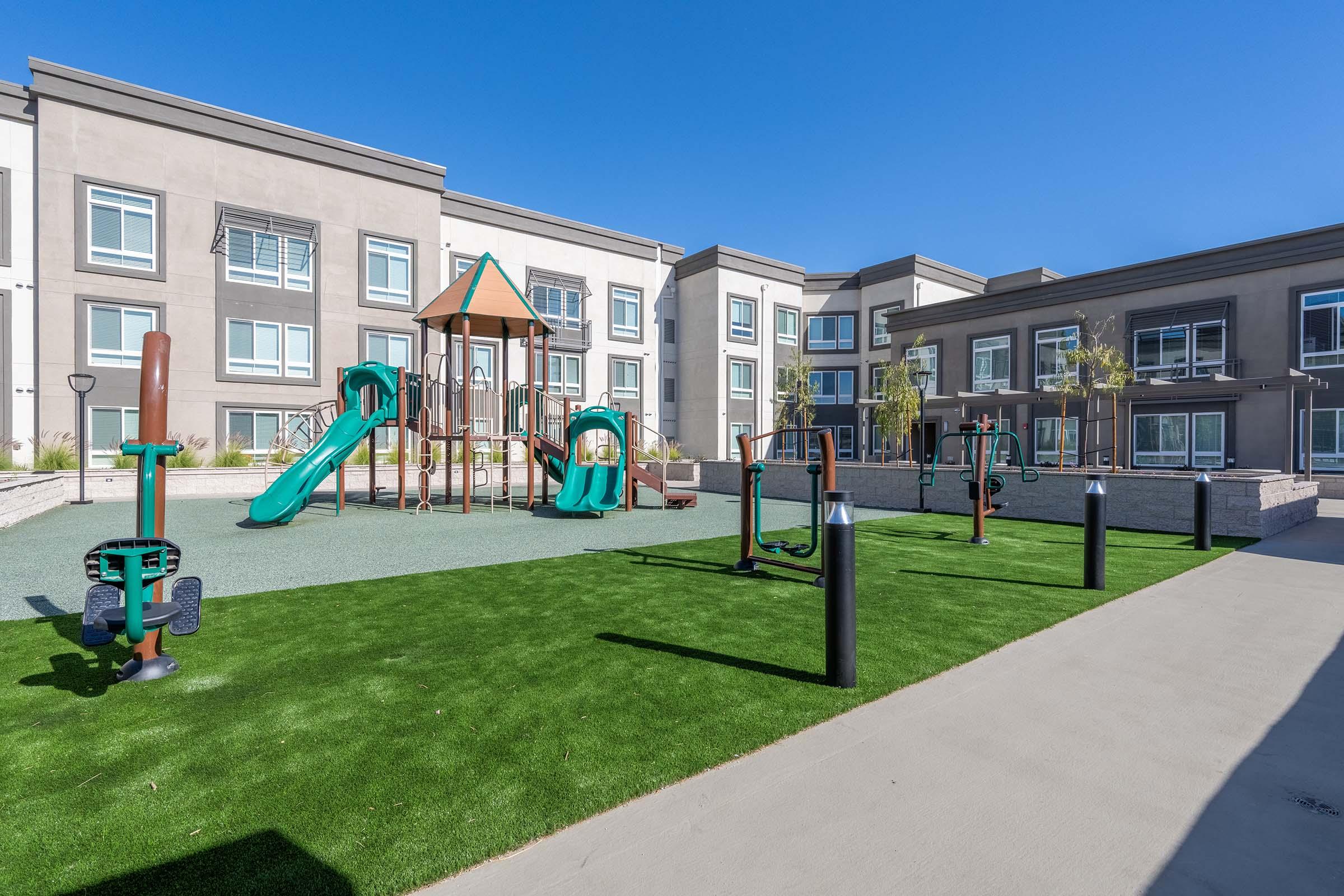 Playground area featuring a wooden play structure with slides, and various fitness equipment on artificial grass. The background shows modern buildings with large windows under a clear blue sky.