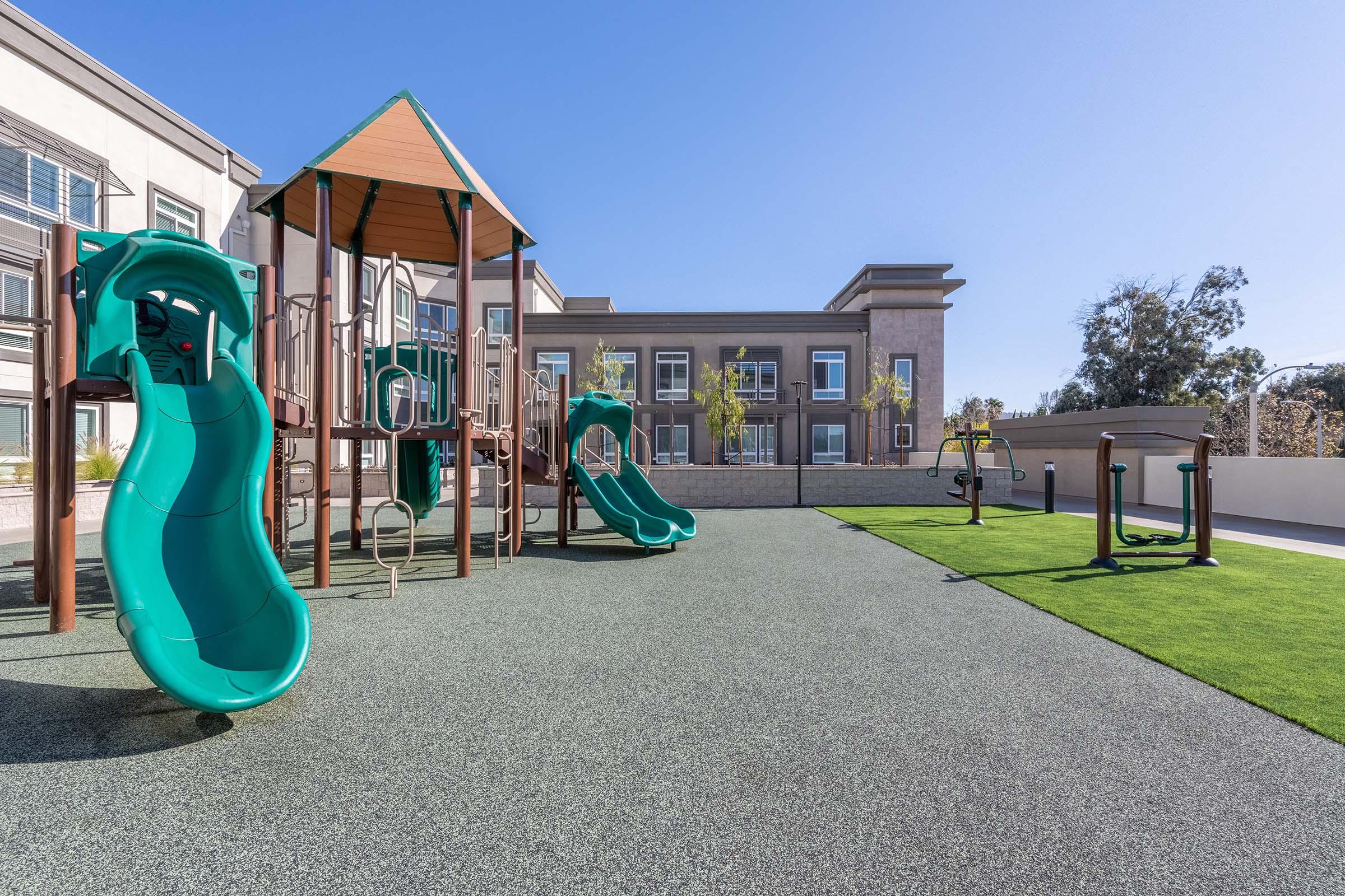 A colorful playground featuring multiple slides and climbing structures, set on soft rubber flooring. In the background, there are modern buildings and landscaping with trees. The area is sunny and inviting, making it ideal for children to play and explore.