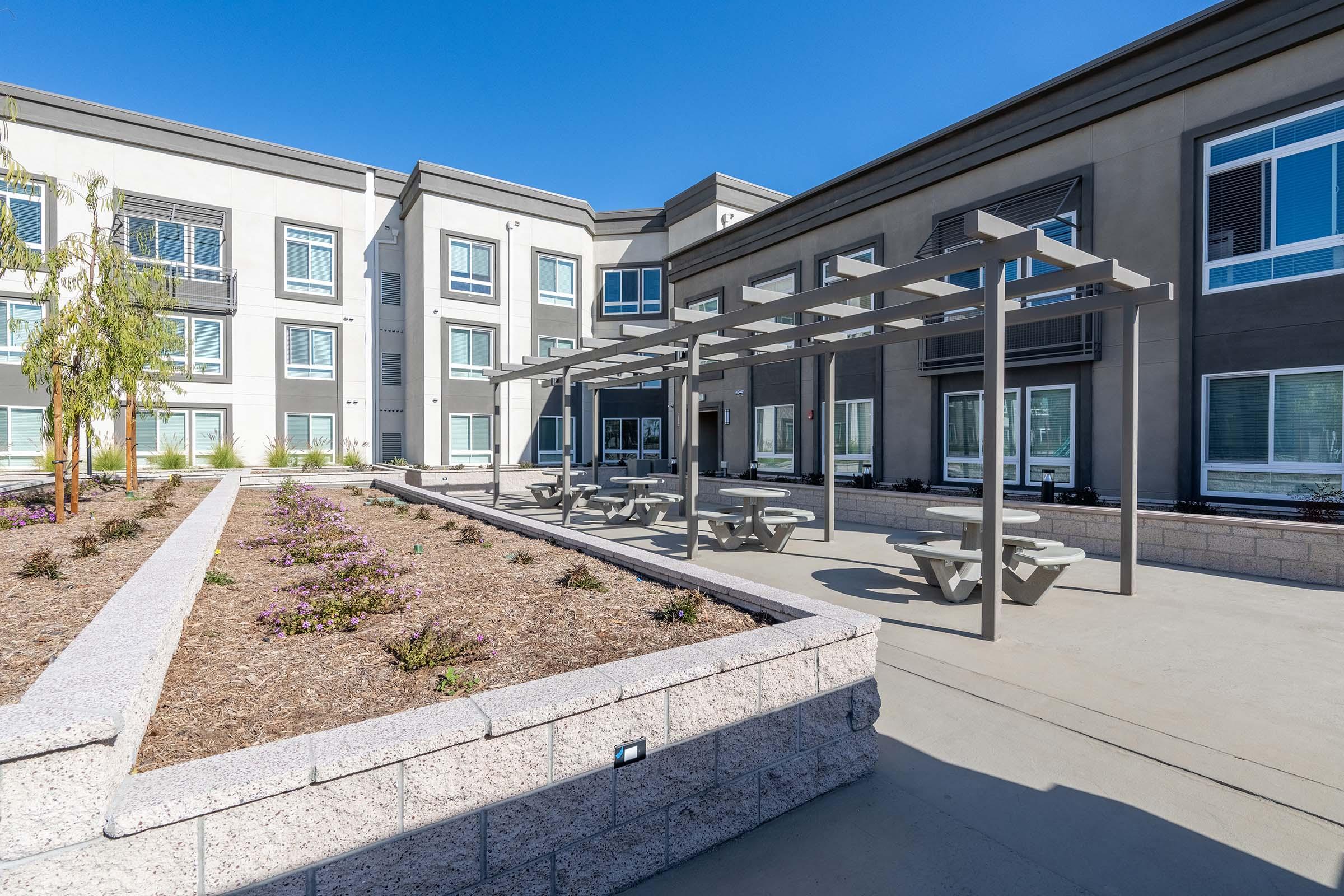 A modern outdoor seating area with picnic tables is surrounded by landscaped flower beds featuring purple blooms. The background shows a multi-story building with large windows, creating a welcoming atmosphere in a sunny, clear blue sky.