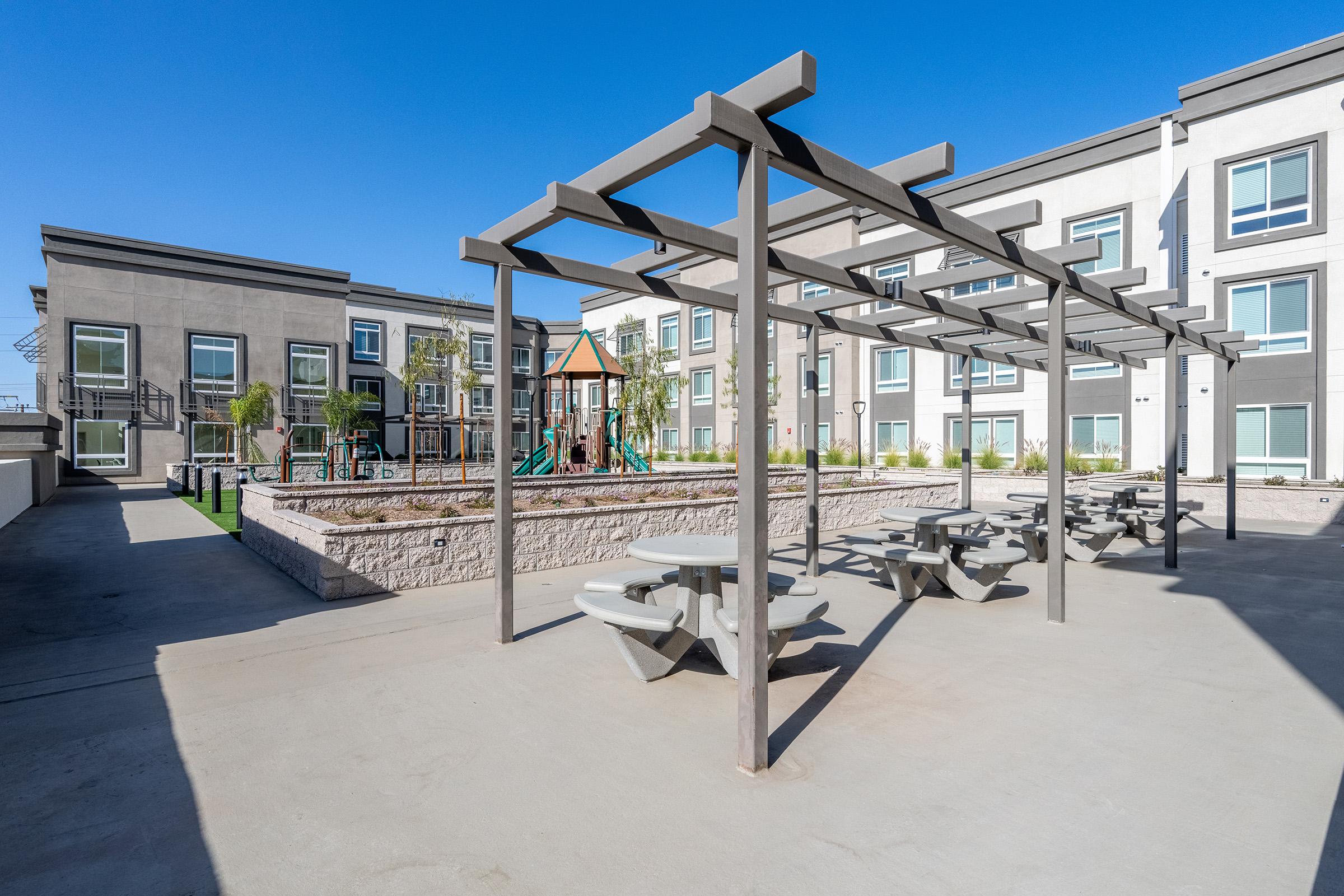A sunny outdoor space featuring a shaded pergola with picnic tables, a children's playground in the background, and a well-maintained landscape. The area is surrounded by modern residential buildings, creating a vibrant community atmosphere. Clear blue sky enhances the inviting environment.