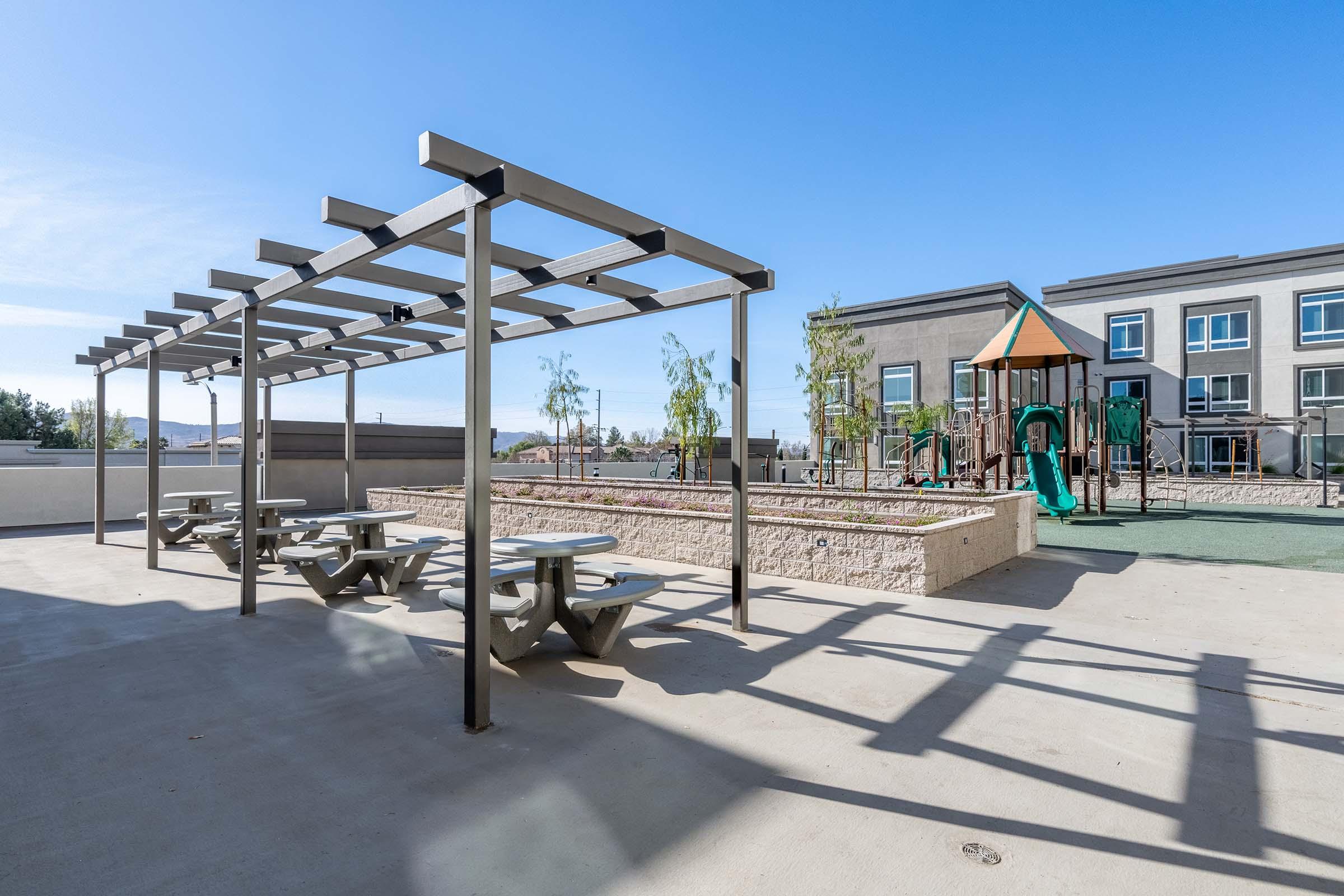 A rooftop playground area featuring a green turf surface, a play structure with slides, and picnic tables underneath a shaded pergola. The background shows modern residential buildings and clear blue skies.