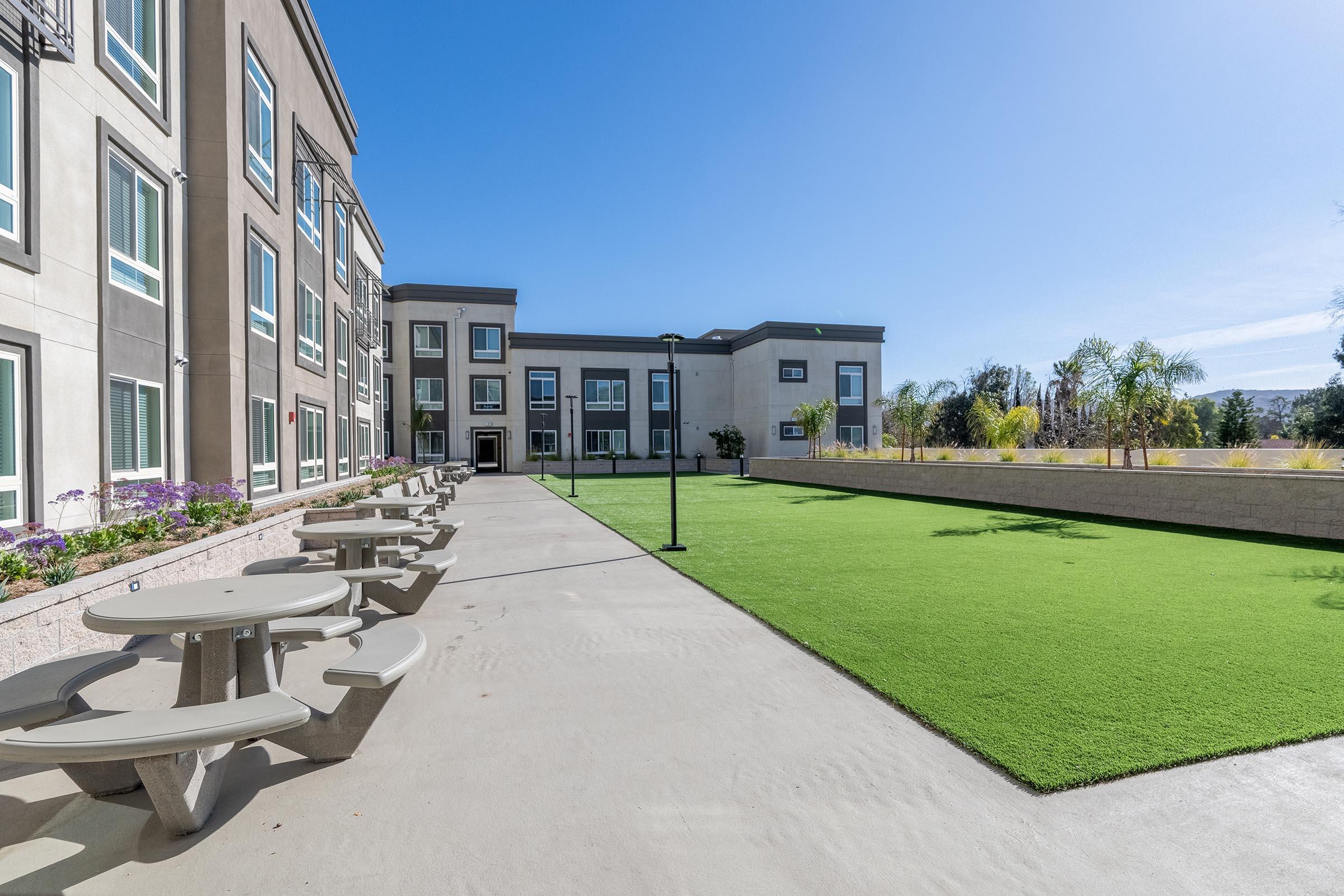 A spacious, well-lit outdoor area featuring a green lawn and multiple picnic tables. The surrounding buildings have large windows and a modern design. The sky is clear and blue, creating a bright and inviting atmosphere for relaxation or social gatherings.