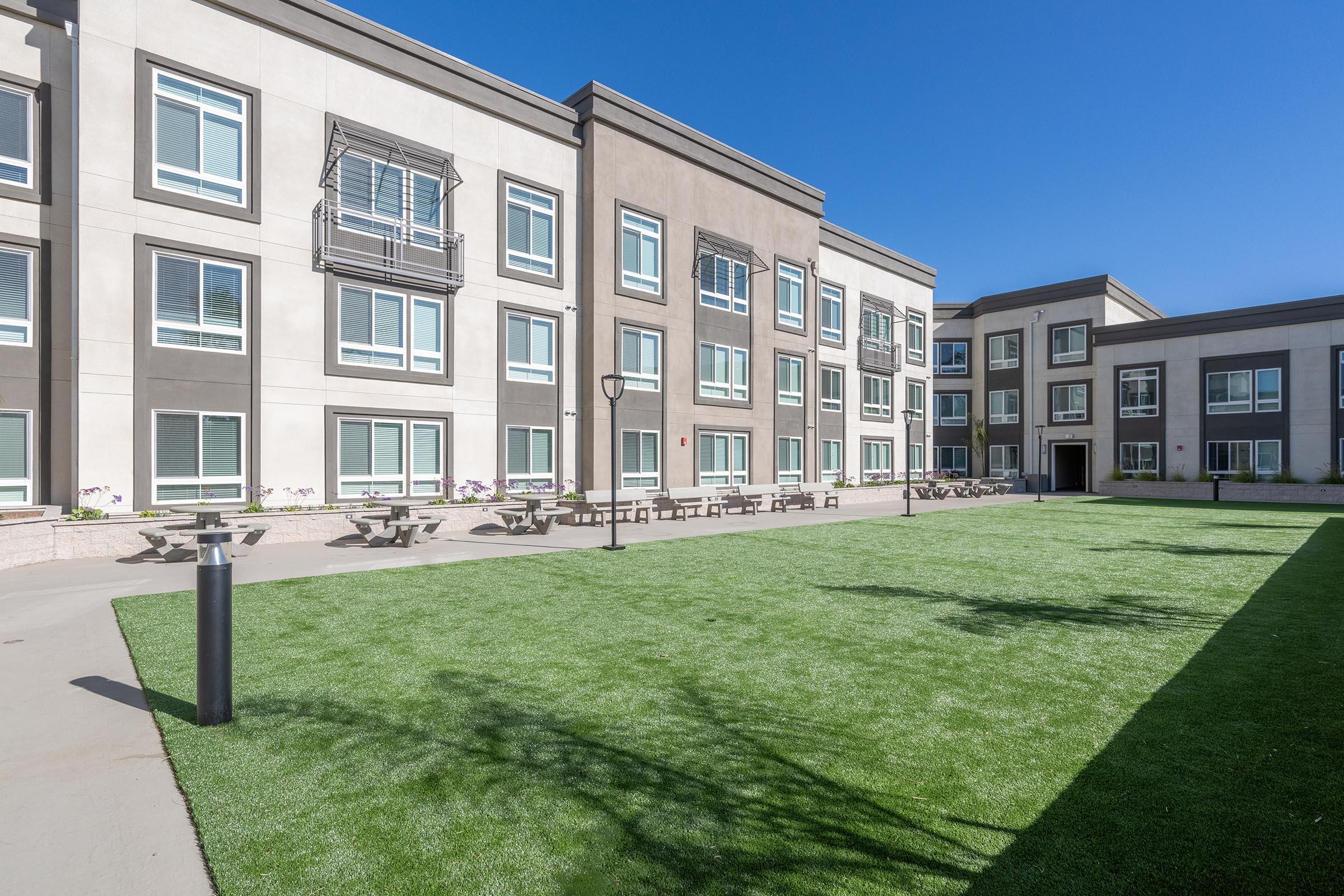 A modern building with multiple windows, featuring a green lawn area in the foreground. Several picnic tables are arranged on the grass, and there are well-maintained pathways. The sky is clear and blue, creating a bright and inviting atmosphere.