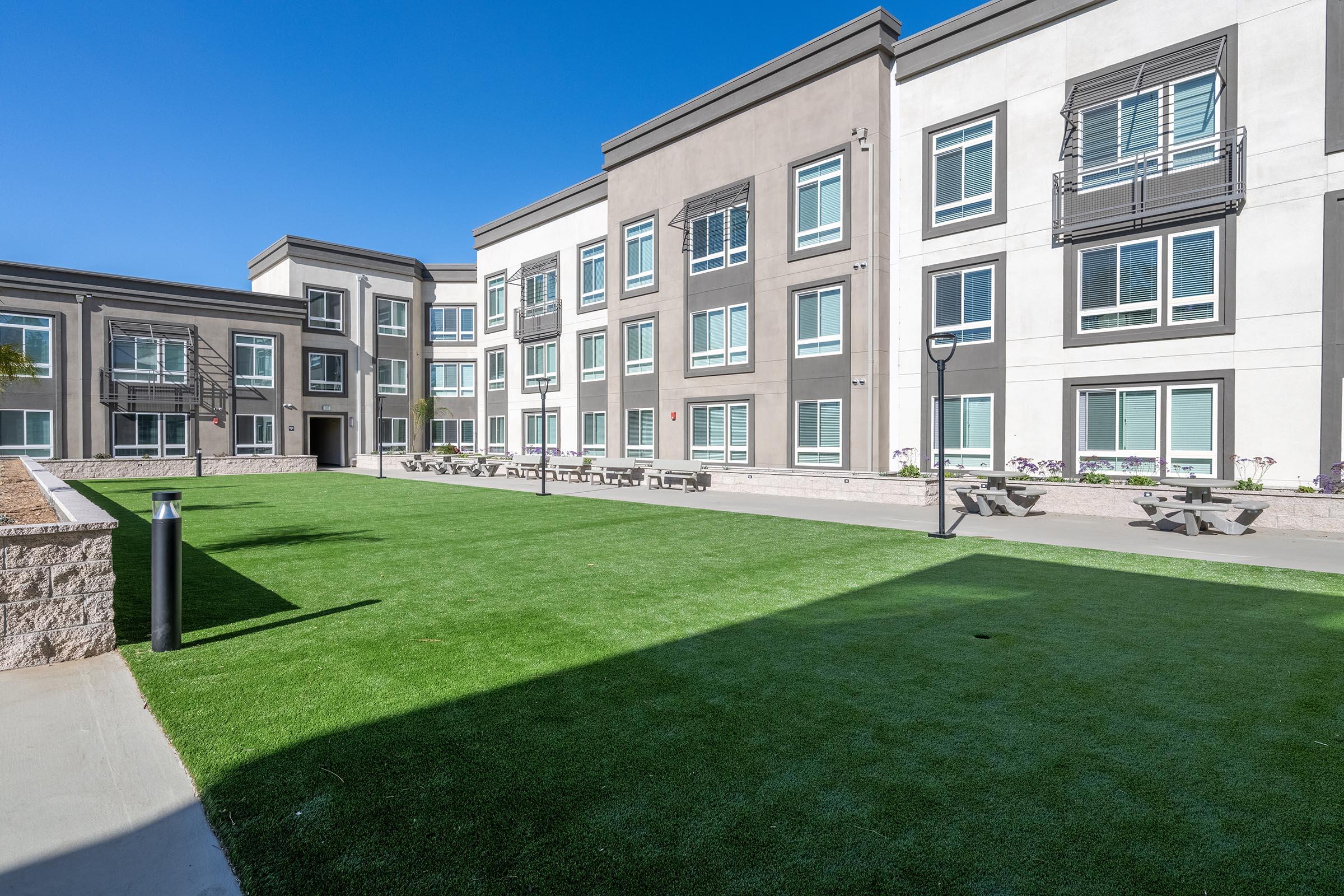 A modern building with multiple floors, featuring large windows and a landscaped courtyard. The courtyard is covered in artificial grass, and there are picnic tables for outdoor seating. The clear blue sky adds a bright atmosphere to the scene.