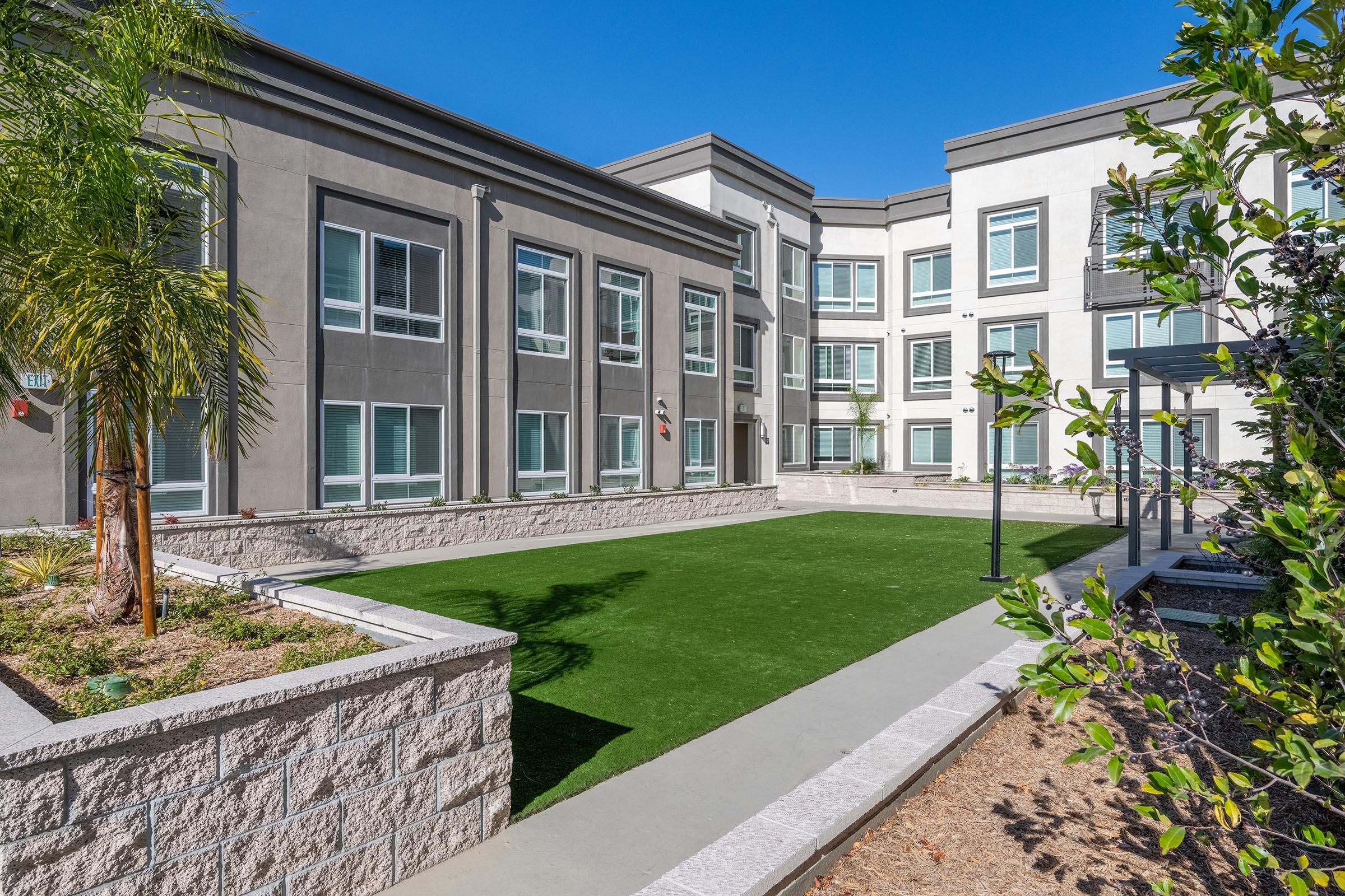 View of a modern apartment complex featuring a landscaped courtyard with artificial grass and palm trees. The building has multiple large windows and a sleek architectural design. Clear blue sky above adds to the bright atmosphere.