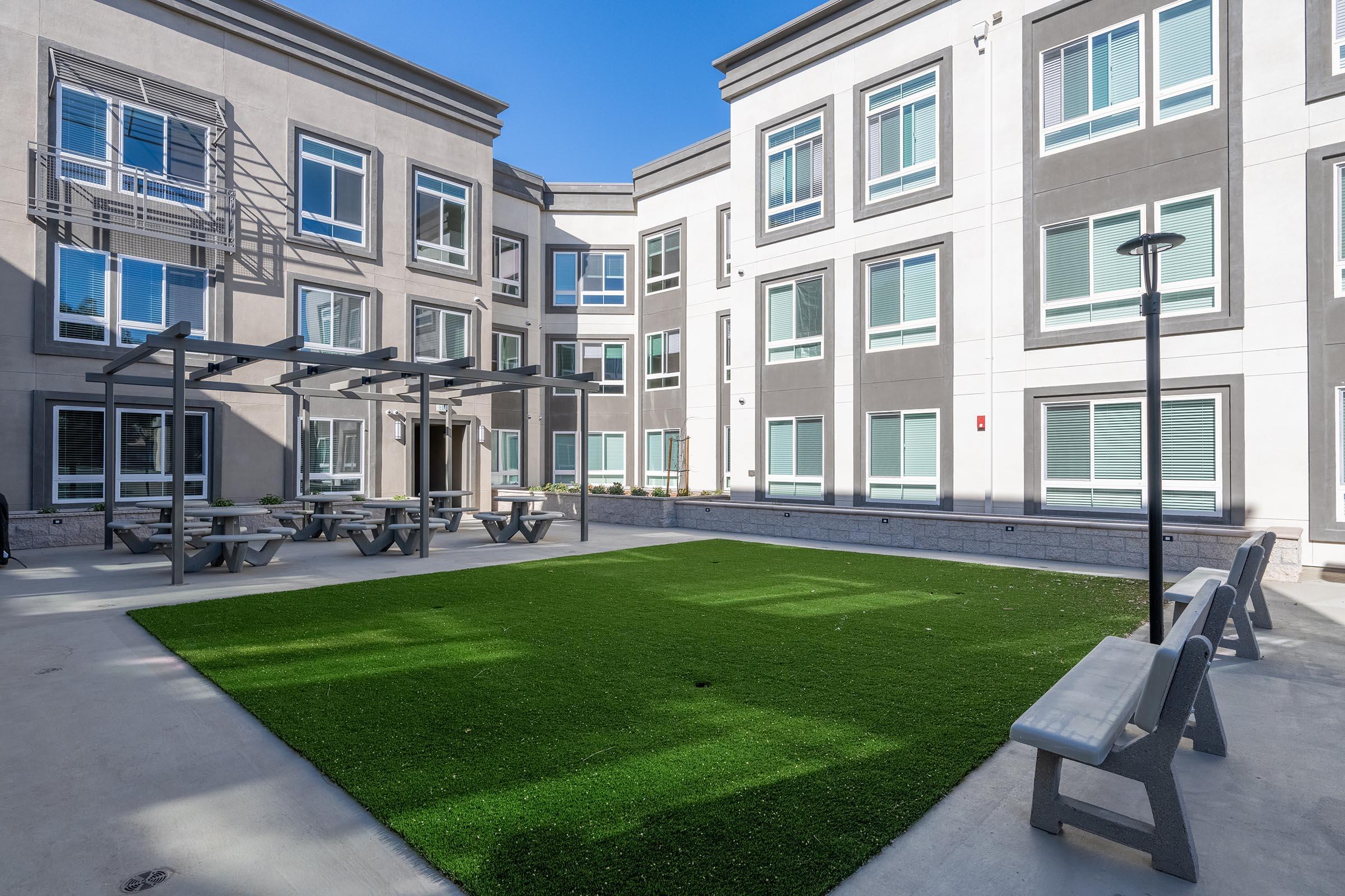 A courtyard area featuring artificial grass, benches, and picnic tables, surrounded by modern multi-story buildings with rectangular windows. The clear blue sky enhances the bright and open atmosphere of the space.