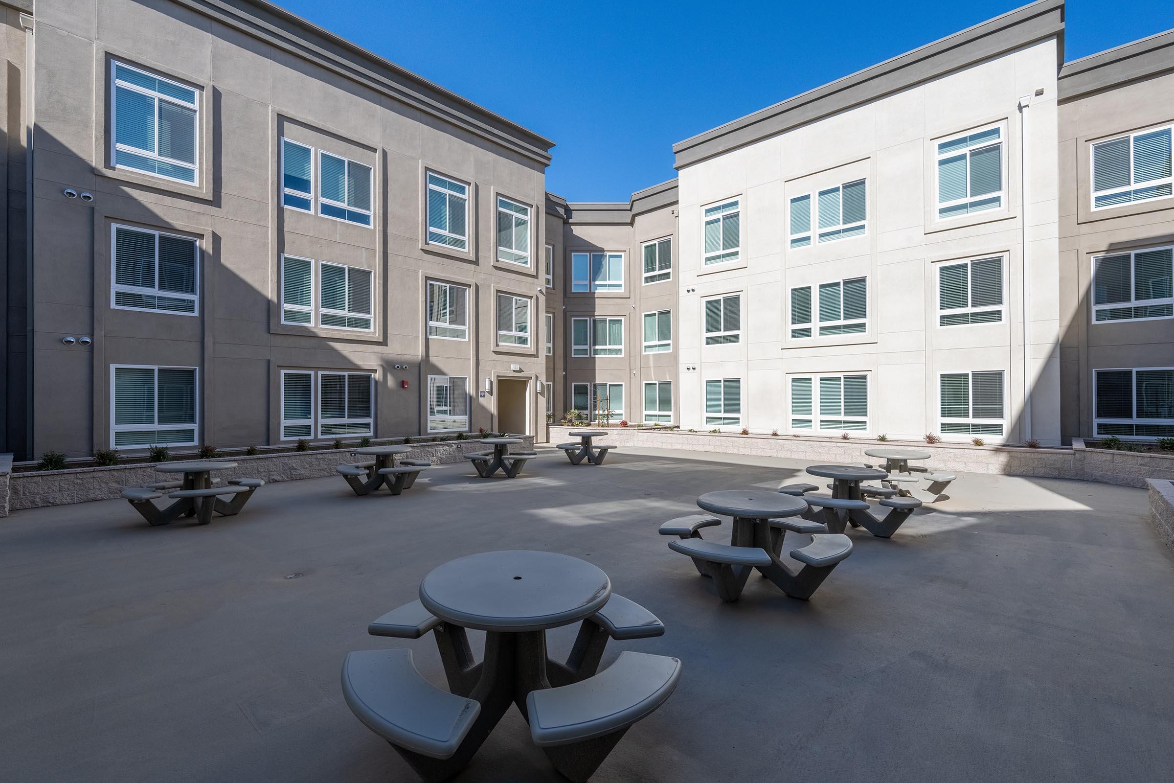 A courtyard surrounded by modern buildings featuring multiple picnic tables and benches. The surface is paved and there are several large windows visible on the buildings, which have a contemporary design. The sky is clear and blue, creating a bright atmosphere.