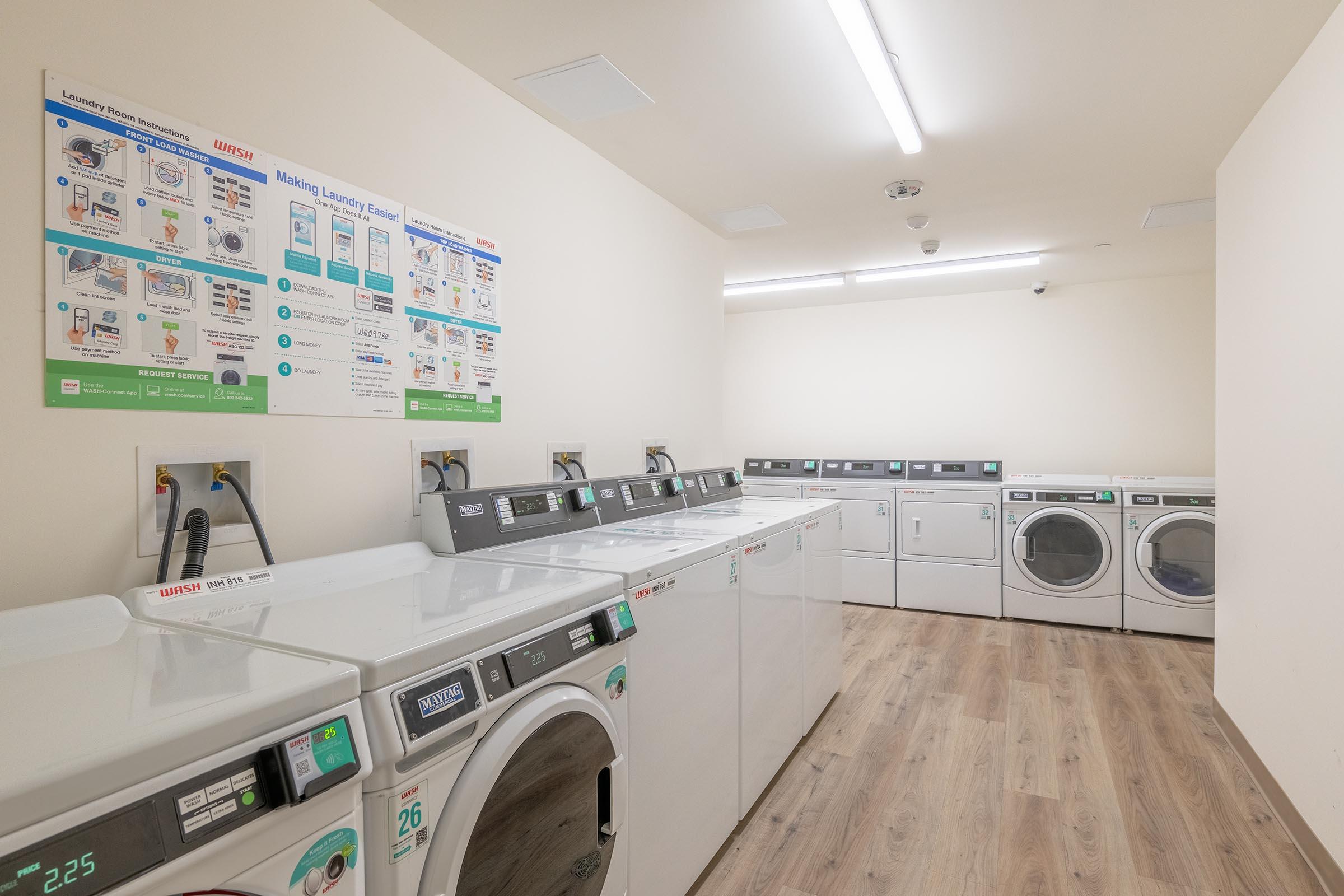 A clean laundry room featuring multiple washing machines and dryers arranged in two rows. The walls display instructional posters on how to operate the machines. The floor is wooden, and the space is well-lit with overhead fluorescent lights.