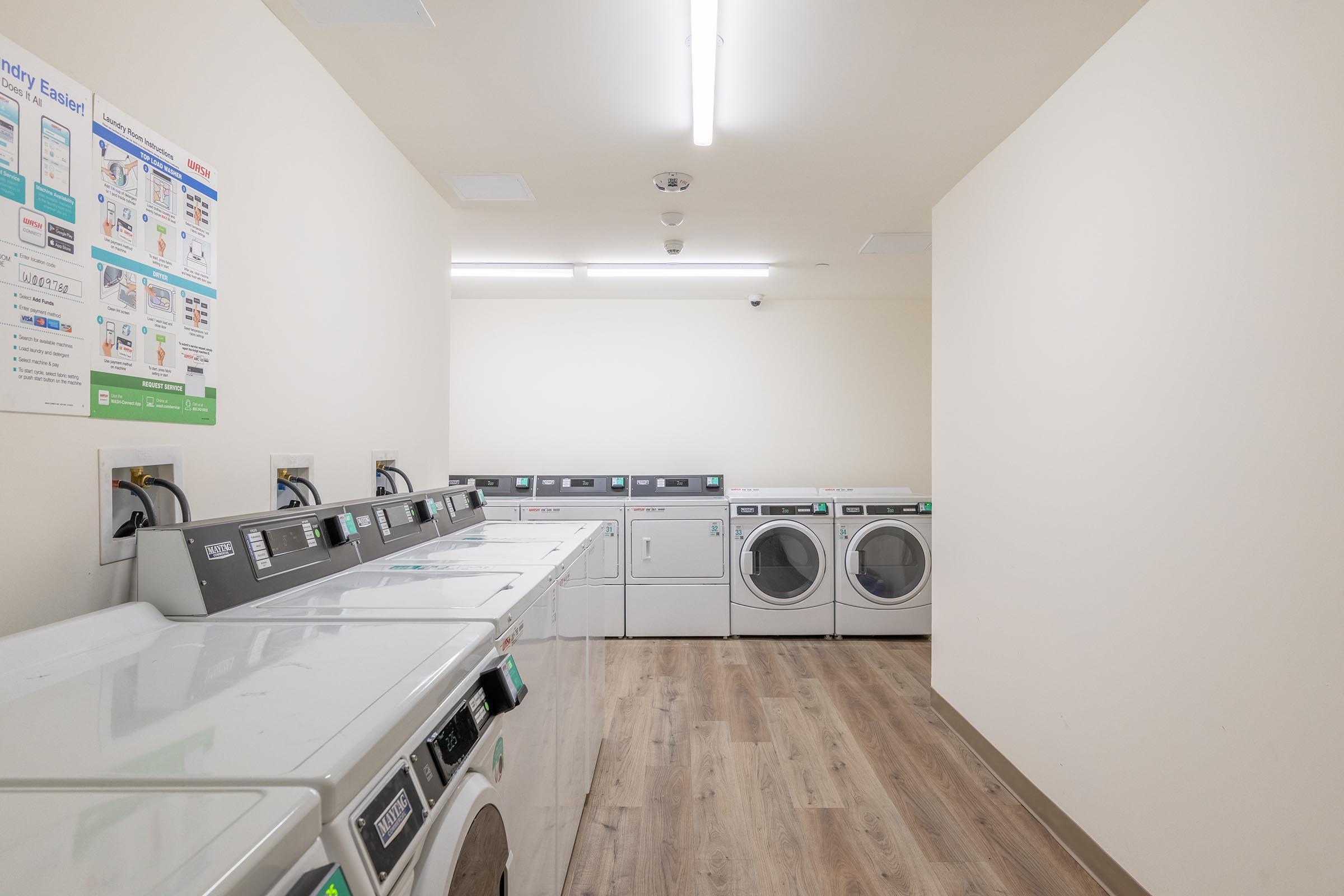 A clean, well-lit laundry room featuring multiple front-loading washing machines and dryers. The machines are lined up against the wall, with a light-colored, wooden floor. A bulletin board with laundry instructions is visible on the left side. The overall ambiance is modern and tidy.