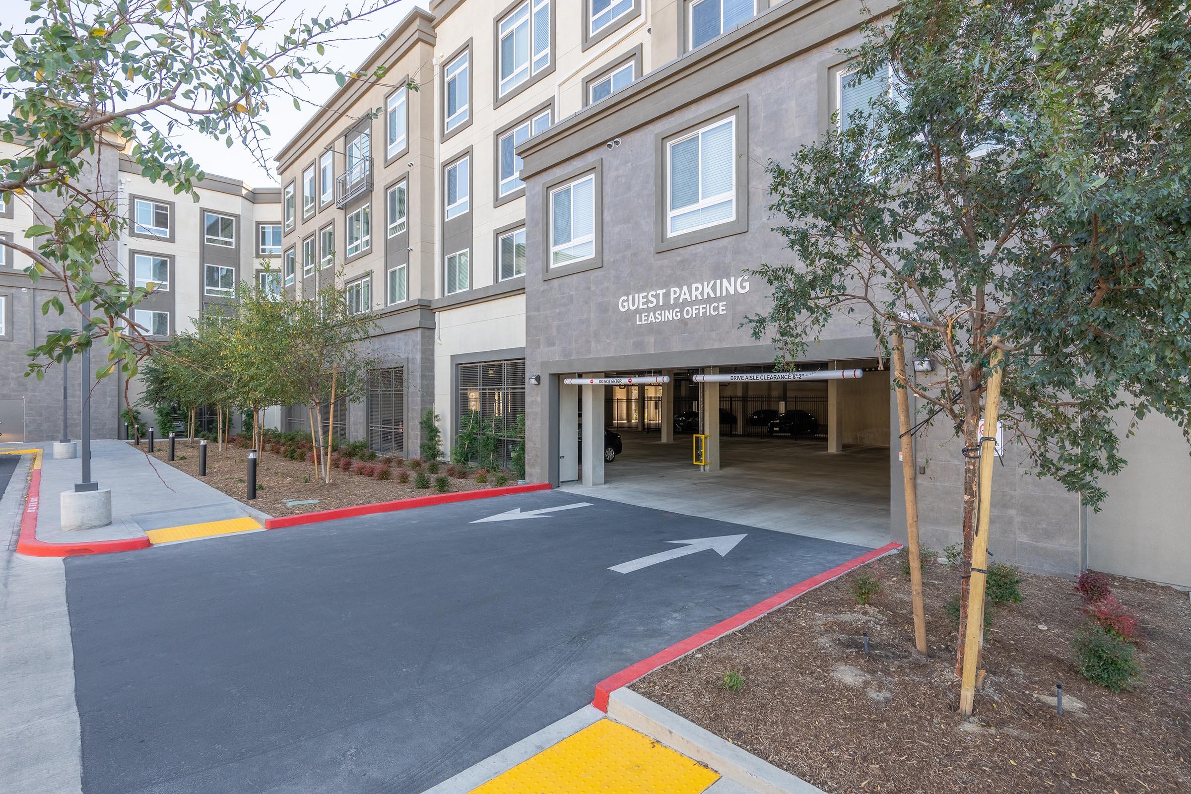 A view of a guest parking area in front of a modern building. The entrance features a sign indicating "GUEST PARKING" and "LEASING OFFICE." Surrounding the parking area are small trees and landscaped sections, with a clear driveway leading into the parking garage. The building has multiple floors and residential windows.