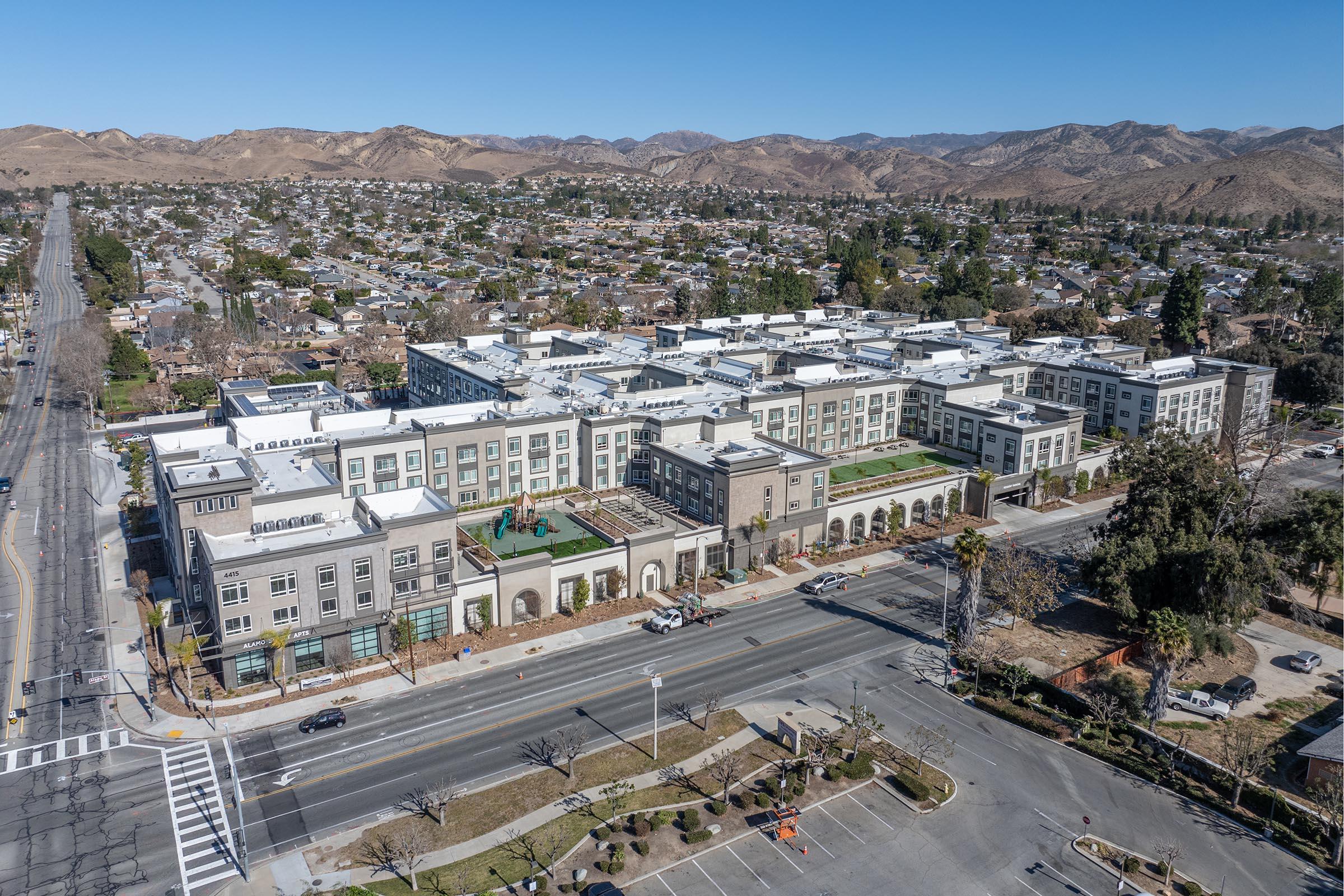 Aerial view of a large modern building complex, featuring several stories with a flat roof. The structure is surrounded by residential neighborhoods and situated near a busy road. In the background, rolling hills and mountains can be seen under a clear blue sky.