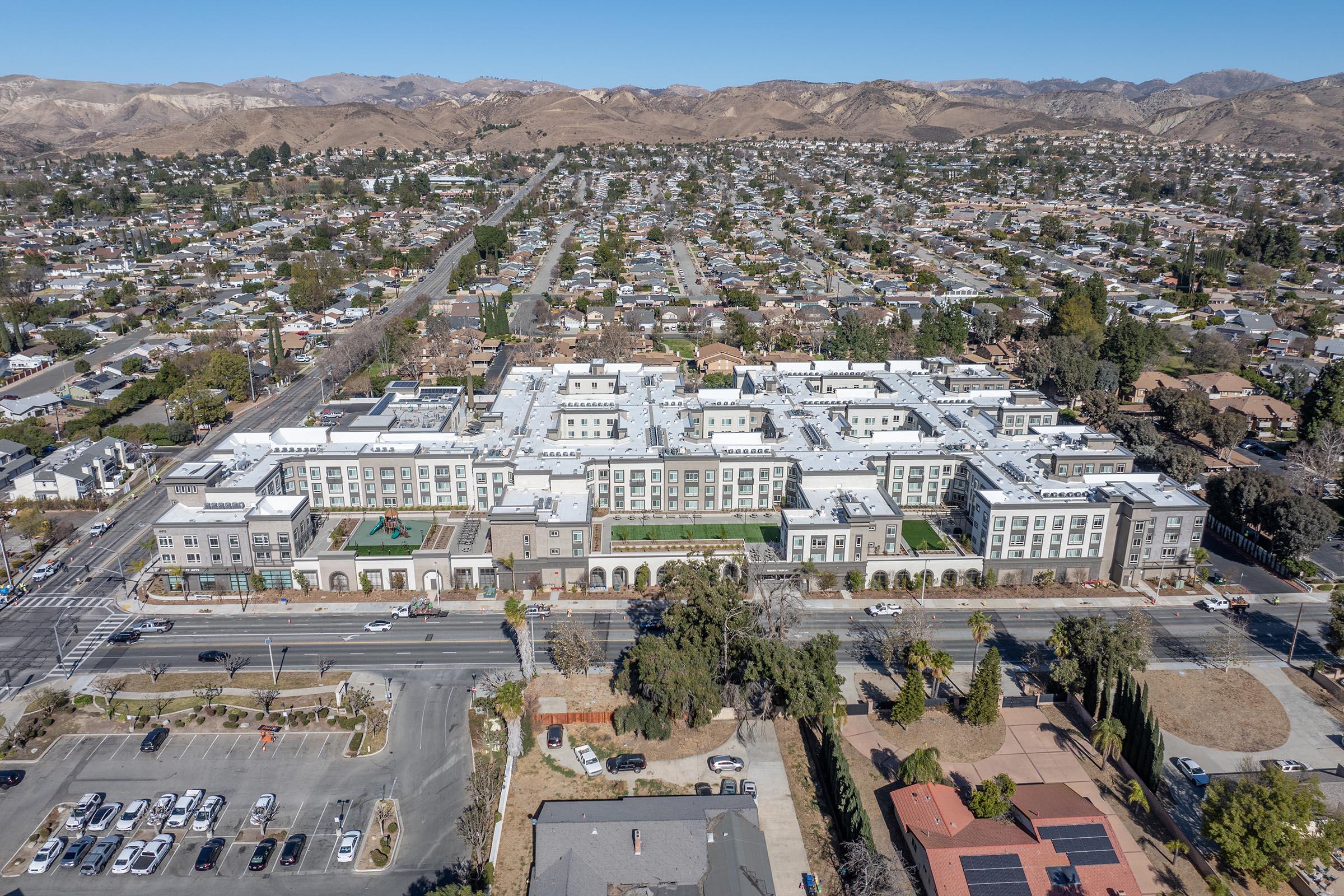 Aerial view of a modern multi-story residential building complex with green spaces and outdoor amenities. Surrounding the complex are a mix of suburban homes and roads, with mountains in the background under a clear blue sky. The area appears well-developed and organized.