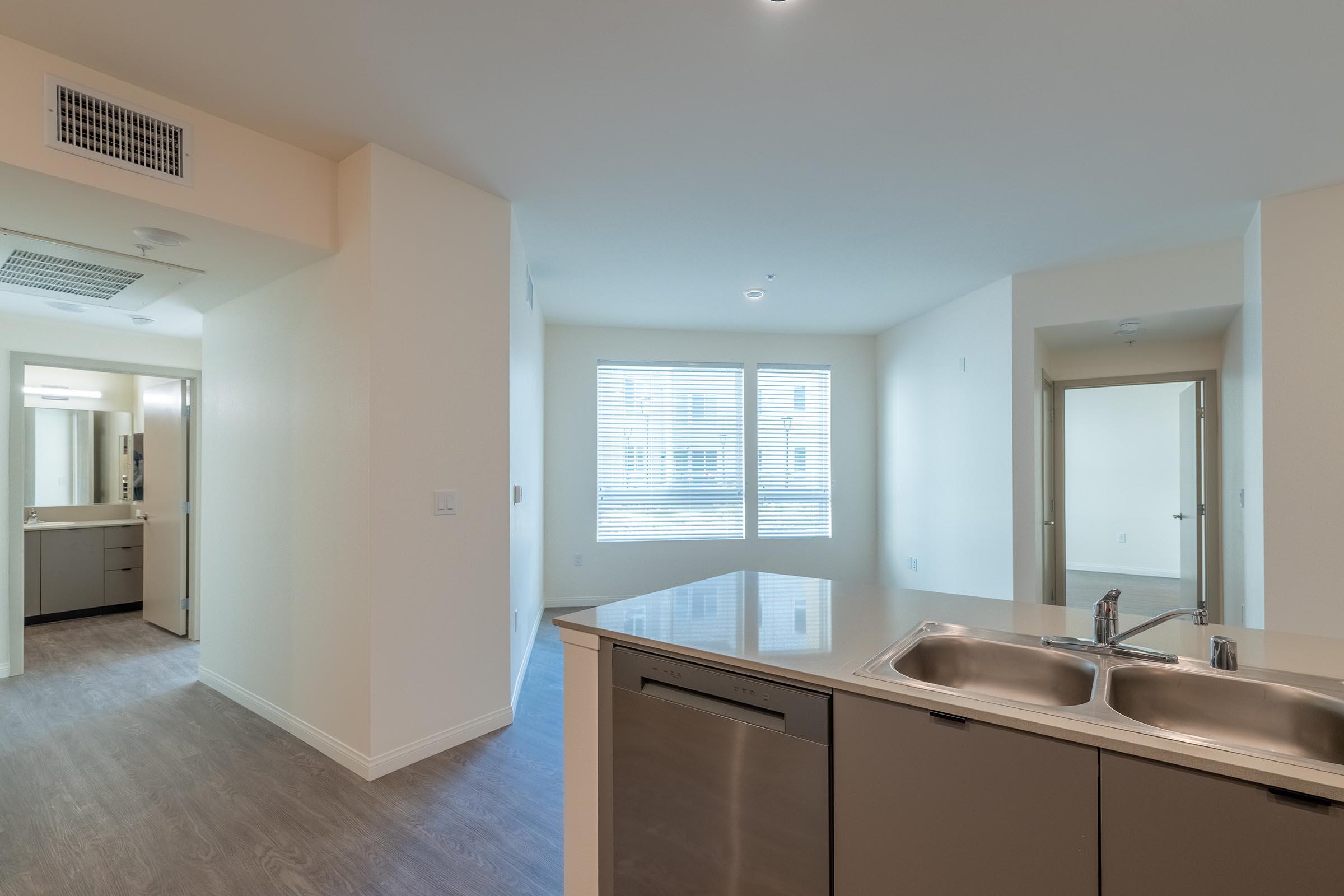 Modern kitchen with a stainless steel sink and dishwasher, open to a bright living area with large windows. Light-colored walls and a seamless floor design lead to a bathroom and closet visible in the background. Natural light fills the space, creating a welcoming atmosphere.