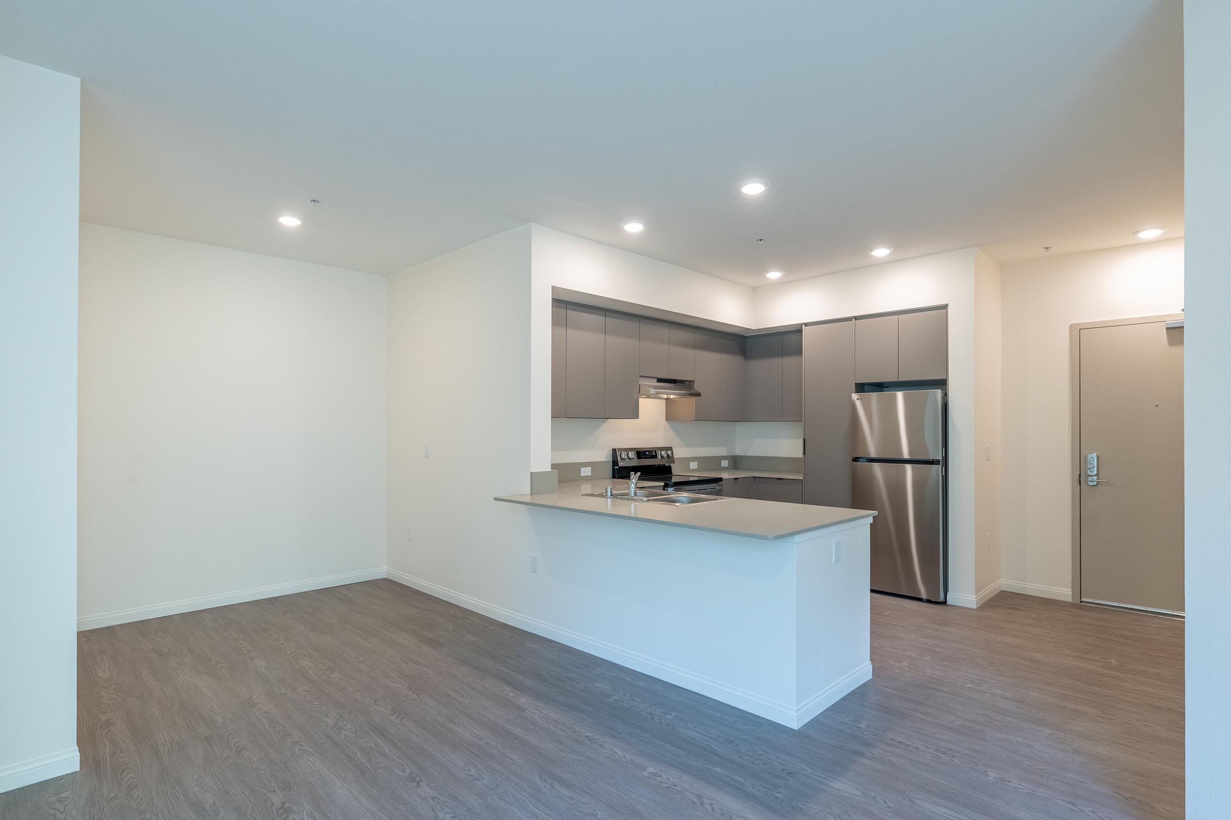A spacious kitchen area featuring modern cabinetry, stainless steel appliances, and a countertop with bar seating. The open layout connects to a well-lit living space with neutral-colored walls and sleek flooring. A front door is visible at the far end, enhancing the inviting atmosphere.