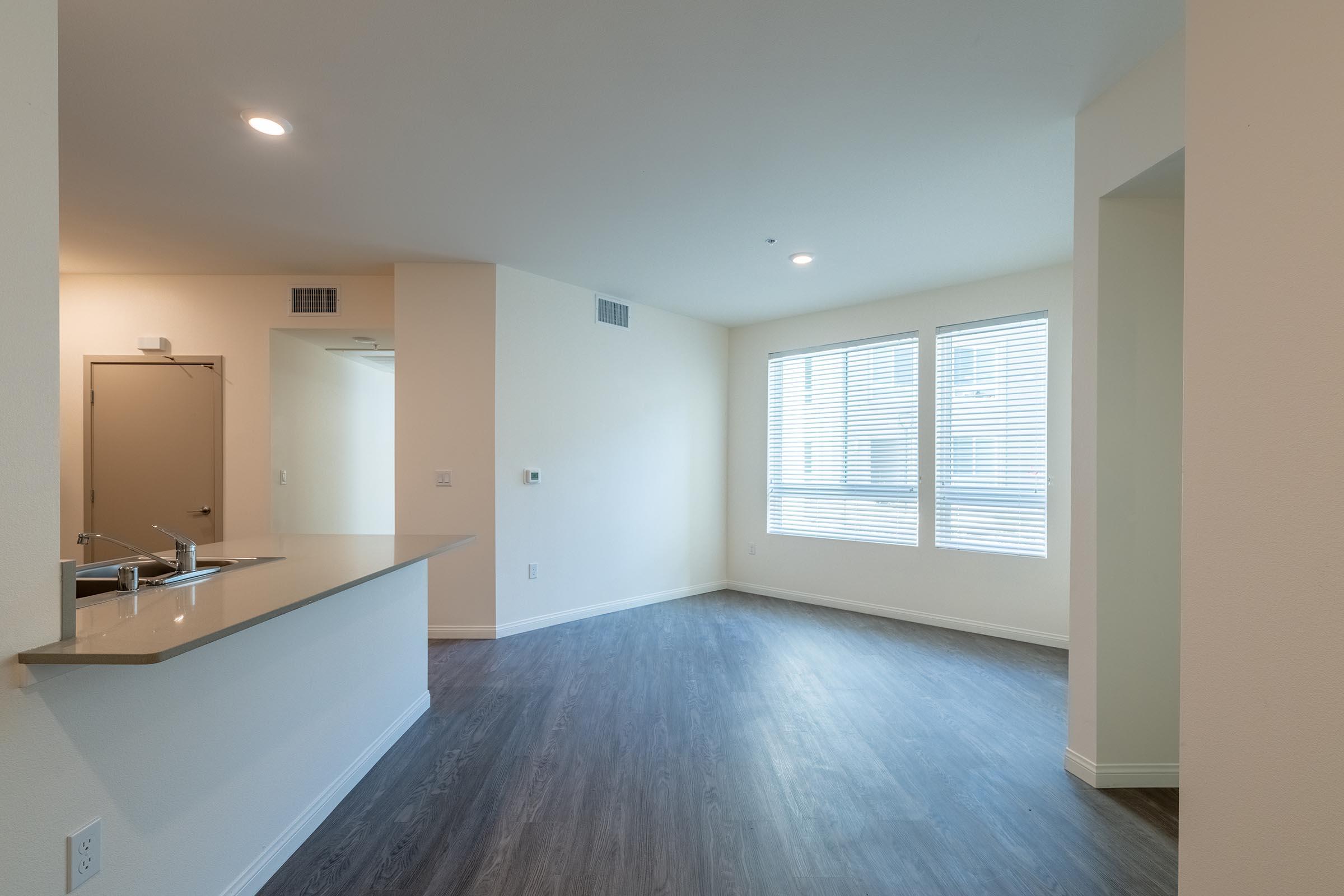 Bright and airy interior of a modern living room with large windows covered by blinds. Features a light-colored wall and a kitchenette with a countertop. Flooring is laminate, creating a warm ambiance. The space appears open and inviting, suggesting a contemporary style.