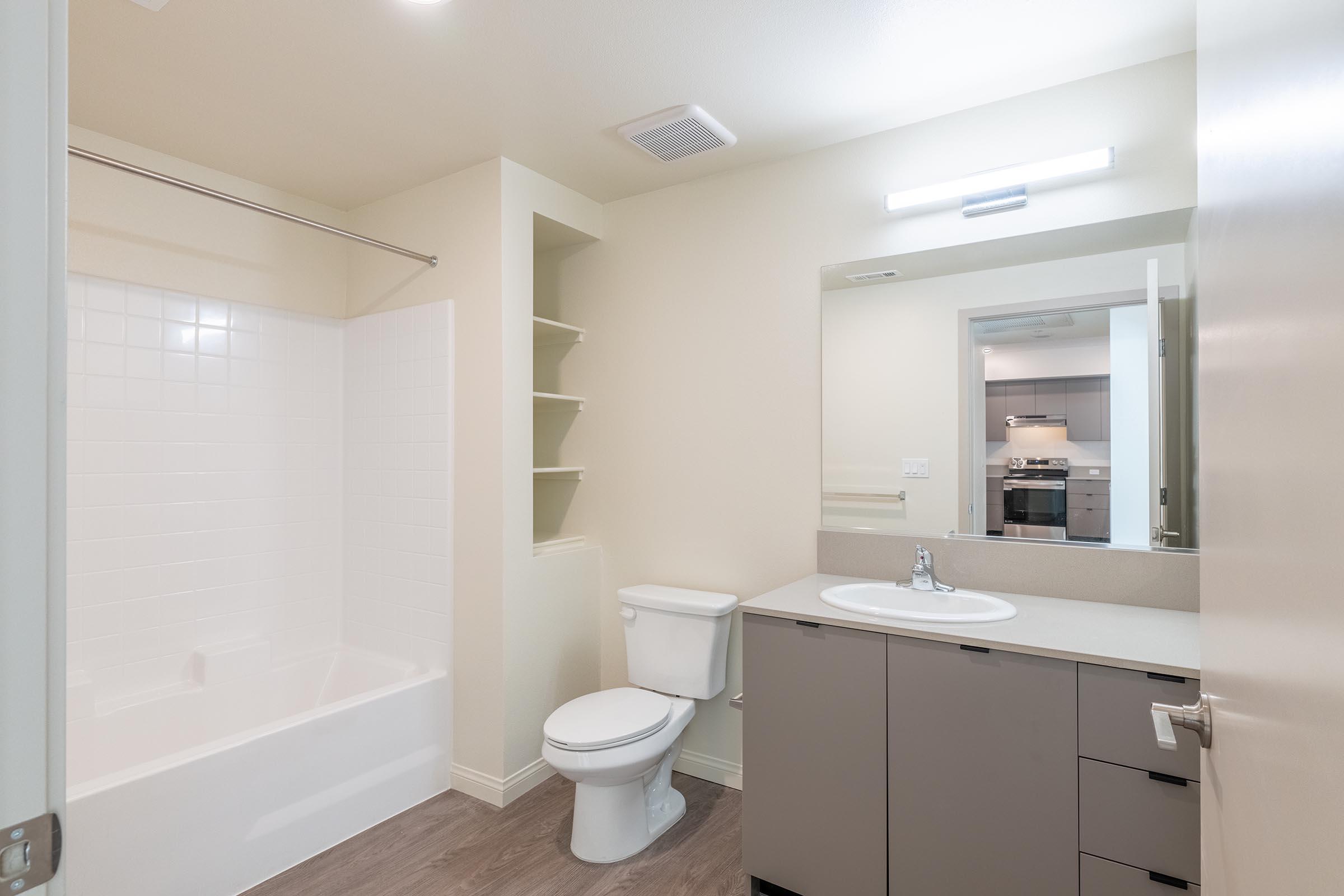 A clean, modern bathroom featuring a white bathtub with a shower, a toilet, a large mirror above a sink with a light fixture, and storage shelves. The walls are light-colored, and the flooring is a light wood finish, contributing to a spacious and contemporary feel.