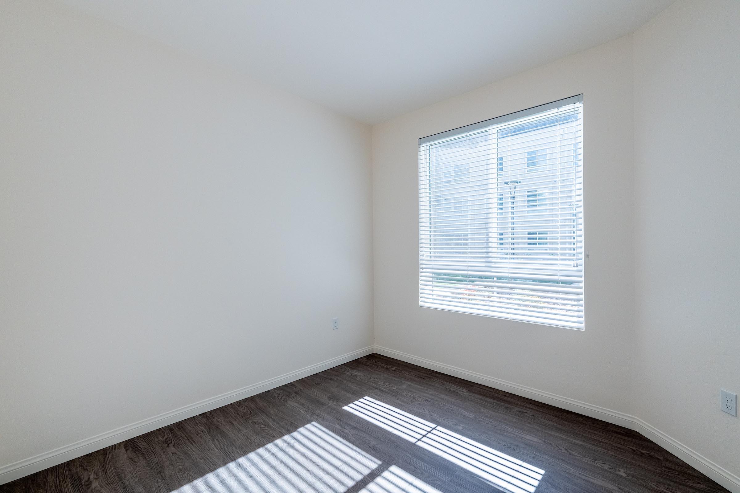 A bright, empty room with light-colored walls and a window covered by white blinds. Light streams in, creating shadows on the brown wooden floor. The room appears spacious and minimalistic, ideal for various uses.
