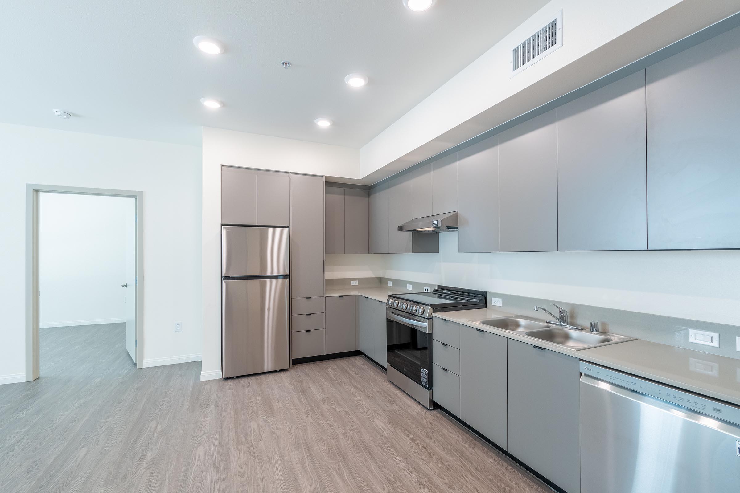 Modern kitchen featuring stainless steel appliances, including a refrigerator, stove, and dishwasher. Sleek grey cabinets and countertops complement the light, airy design. The space has a minimalist aesthetic with ample lighting and an open layout, ideal for contemporary living.