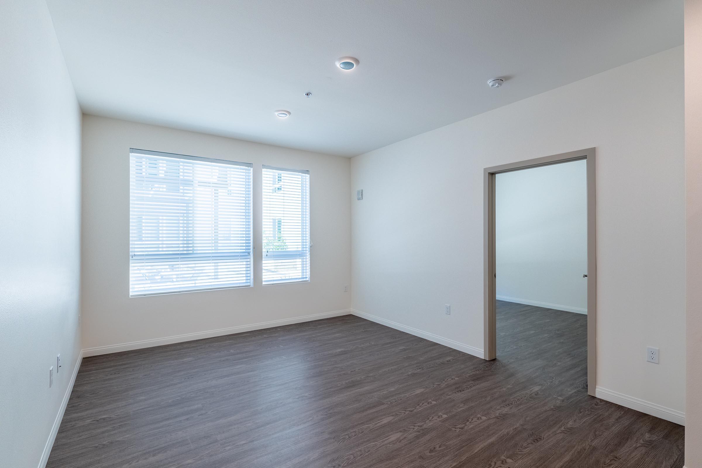 A spacious, empty room with light-colored walls and a medium-toned wooden floor. There are two large windows with white blinds letting in natural light, and a doorway leading to another room. The overall atmosphere is bright and airy, making it suitable for various uses.
