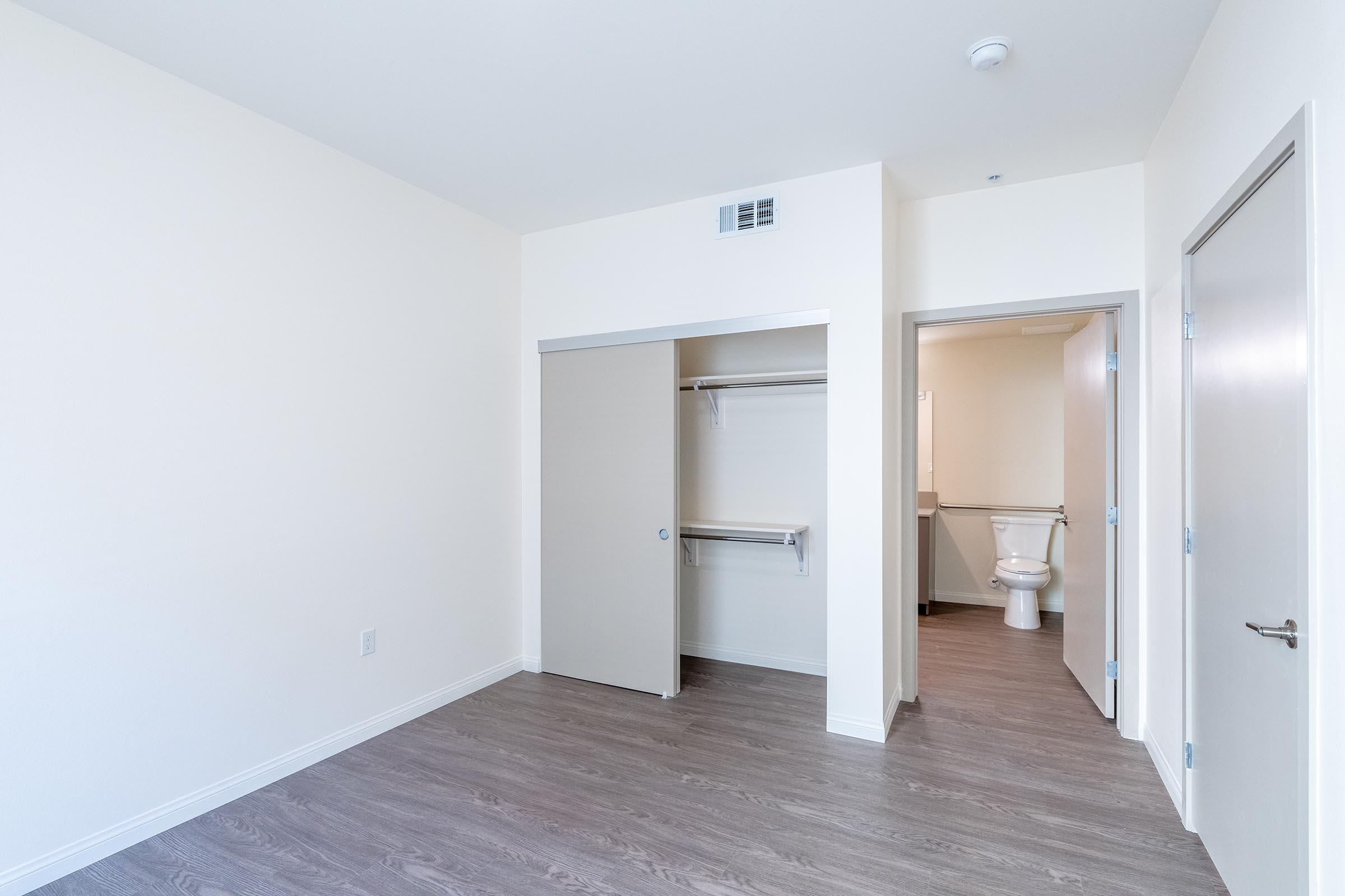 A minimalistic room featuring light-colored walls and a wooden floor. There's a closet with sliding doors on the left, and a small bathroom doorway on the right, partially visible. The space is well-lit and appears clean and modern, suitable for various uses such as a bedroom or office.