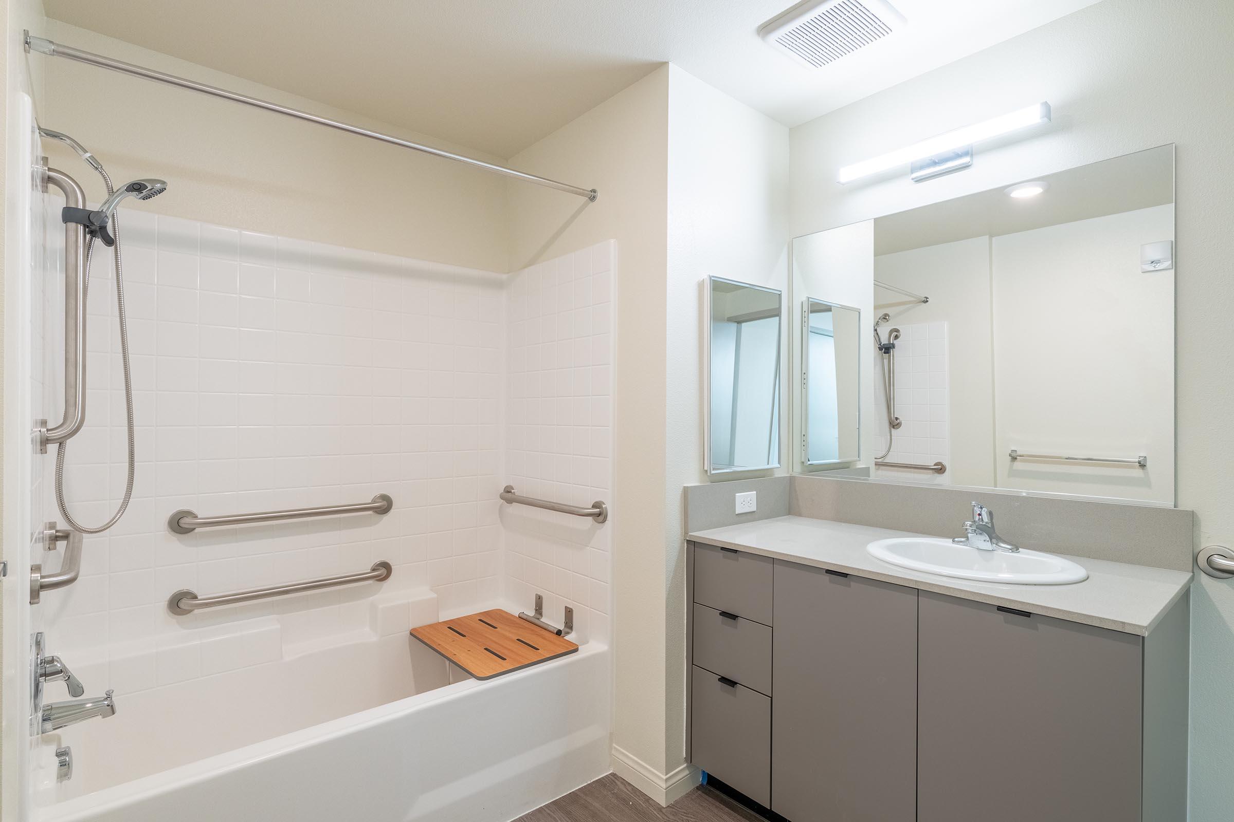A clean and modern bathroom with a white tiled bathtub featuring grab bars, a wooden bath board, a large mirror above a sink with a faucet, and sleek gray cabinetry. The space is well-lit with a ceiling light and has a neutral color scheme, creating a spacious and inviting atmosphere.