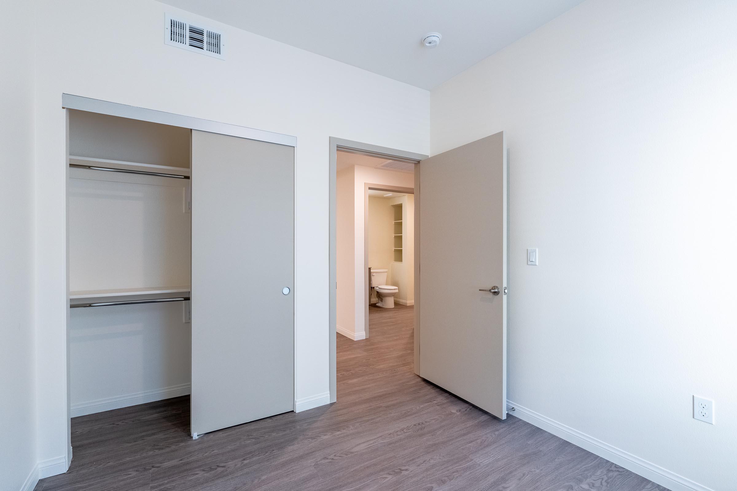A well-lit room with light-colored walls and flooring. There is a closet with sliding doors to the left and an open doorway leading to a bathroom in the background. The bathroom is not fully visible but includes a glimpse of a white toilet and shelves.