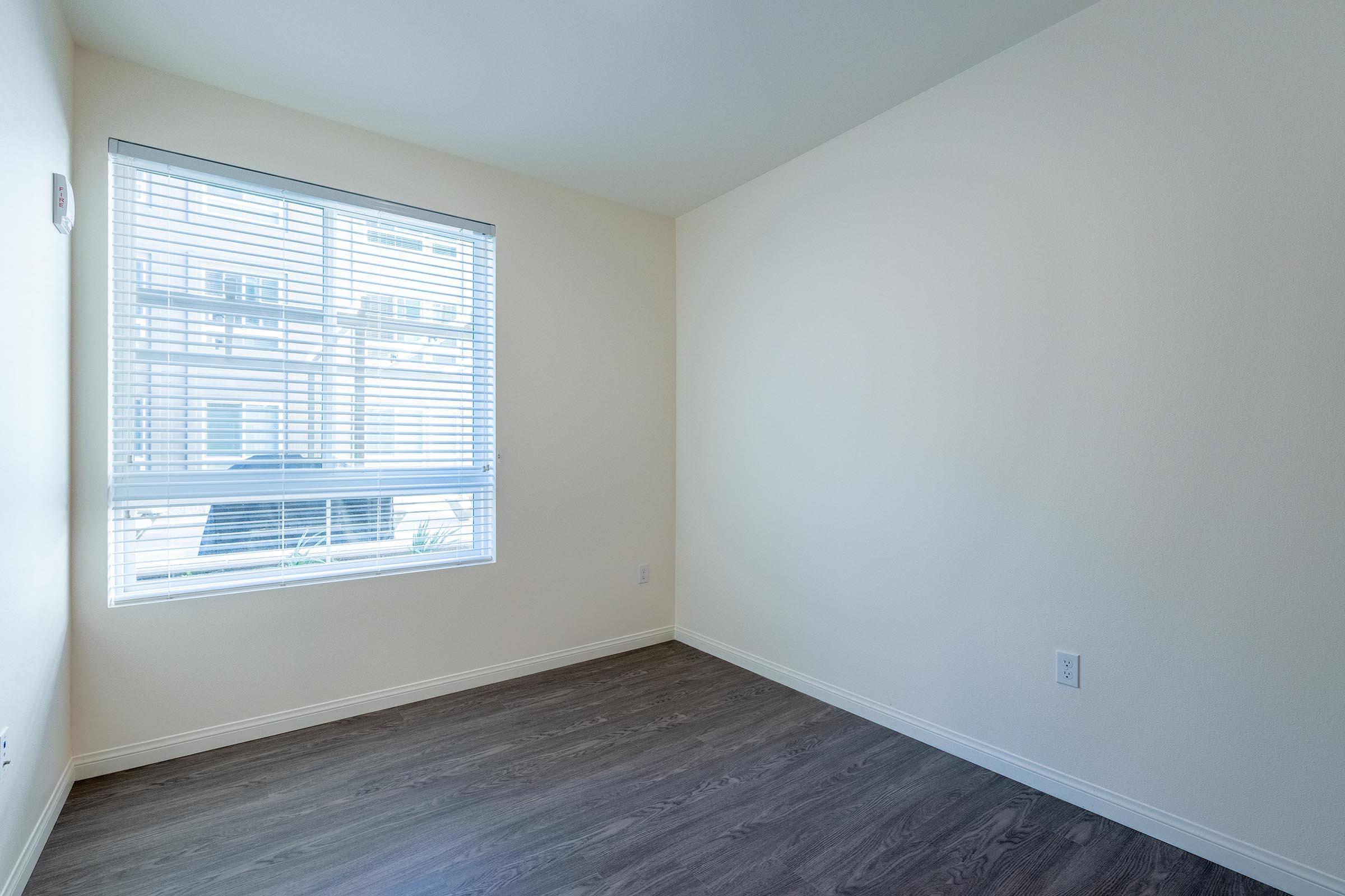 Empty room with light-colored walls and a window featuring horizontal blinds. The floor is dark wood, and there is a small wall thermostat near the window. The space is tidy and unadorned, creating a minimalist atmosphere.