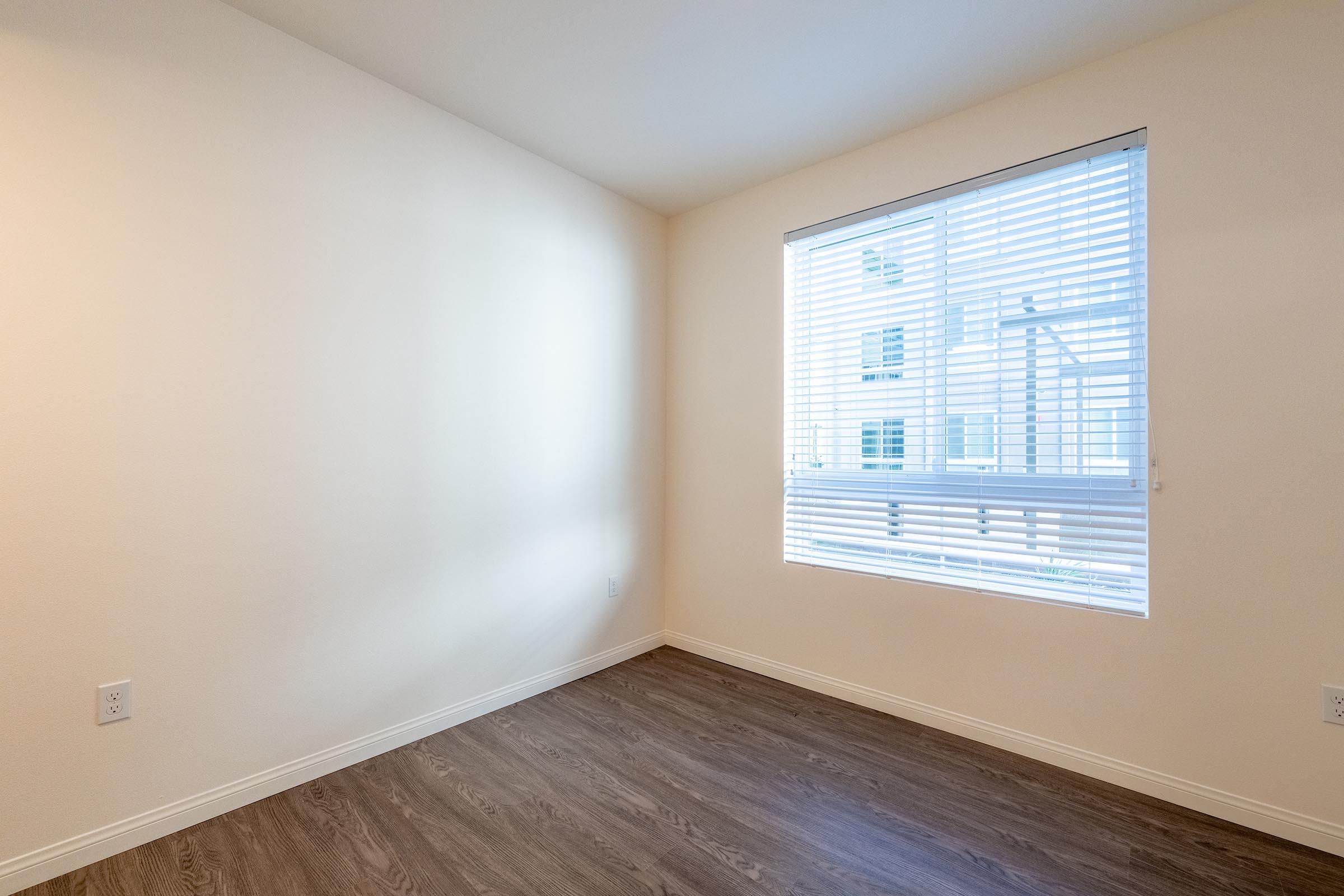 A sparsely furnished room with light-colored walls and a large window covered by white blinds. The floor is a light wood finish, and the overall ambiance is bright and airy, suggesting a new or recently painted space. There are no furnishings or decorations present.
