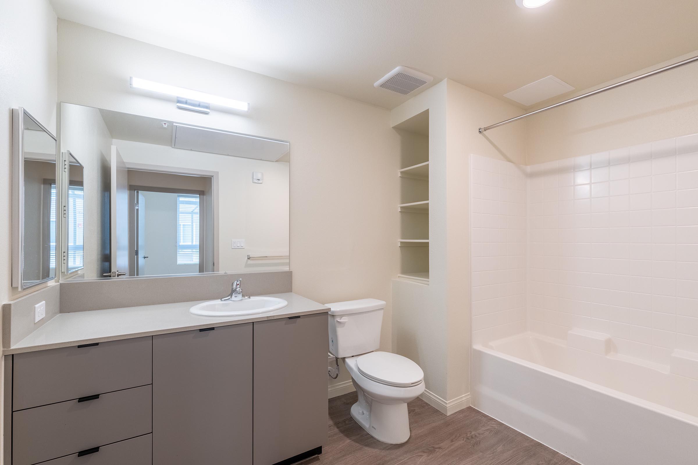 A modern bathroom featuring a white bathtub with shower, a light-colored countertop with a sink, two rectangular mirrors above the sink, a toilet, and open shelving. The walls are painted in a neutral tone, and the space is well-lit by overhead lighting.