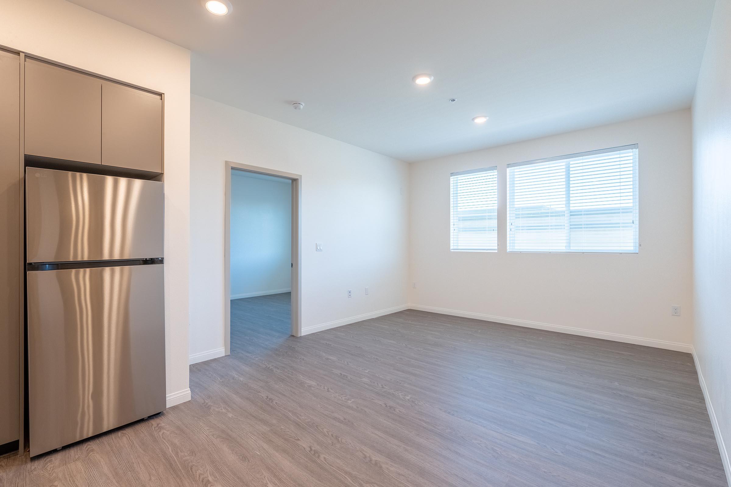 A bright, empty room with light-colored walls and large windows. The space features light gray flooring and a stainless steel refrigerator in one corner. The layout appears modern and uncluttered, providing a blank canvas for decorating. Natural light fills the room, enhancing the open atmosphere.