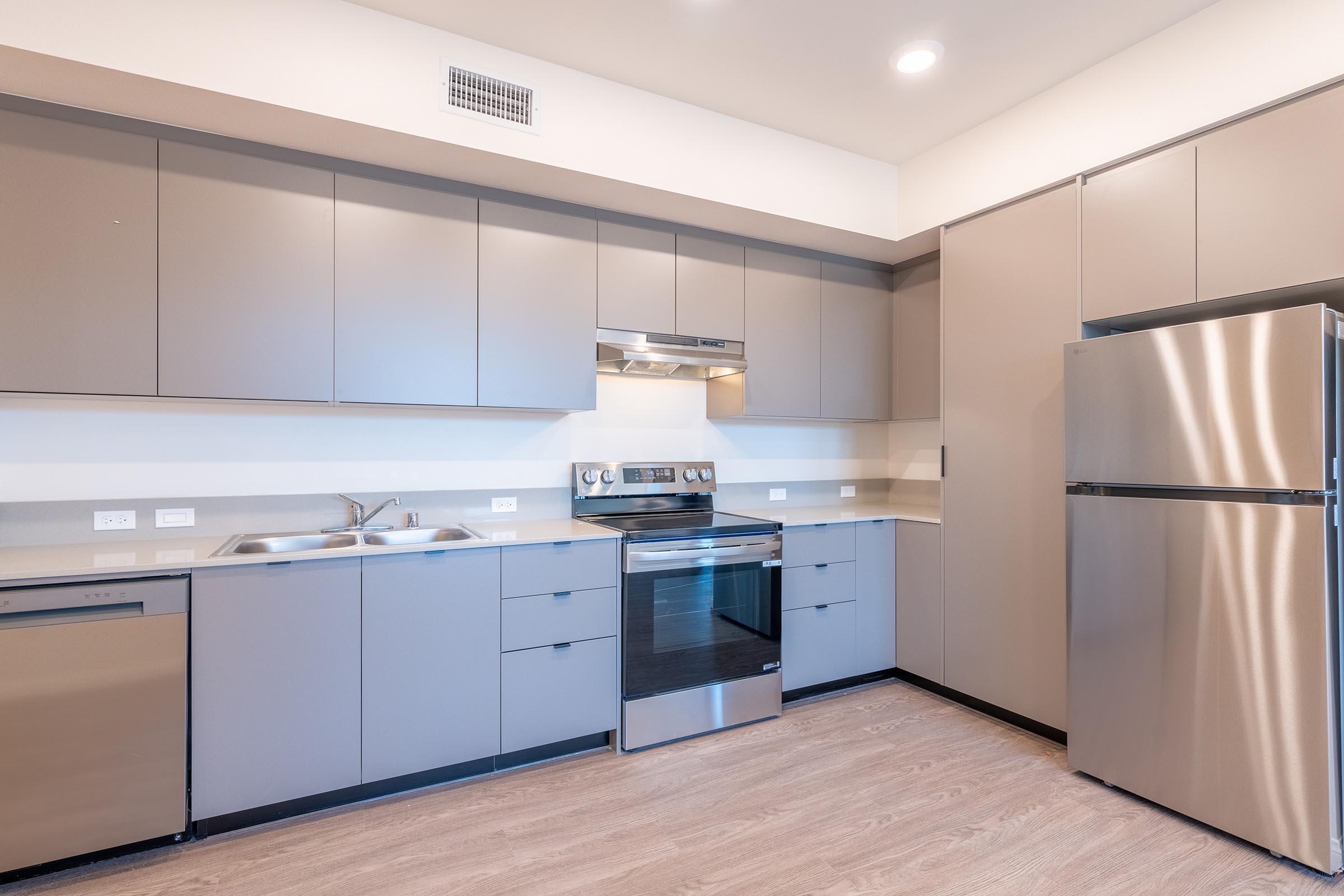 Modern kitchen featuring light gray cabinetry, a stainless steel refrigerator, and an oven with a glass cooktop. The countertop is a smooth surface, and the space has an open layout with ample natural light.