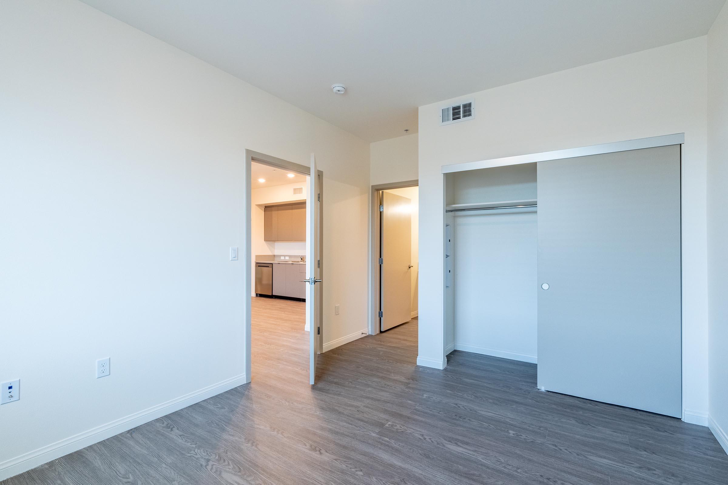 A spacious, empty room featuring light-colored walls and wooden flooring. There's an open door leading to a kitchen area, and a closet with sliding doors on the right. Natural light enters through the doorway, creating a bright, inviting atmosphere.