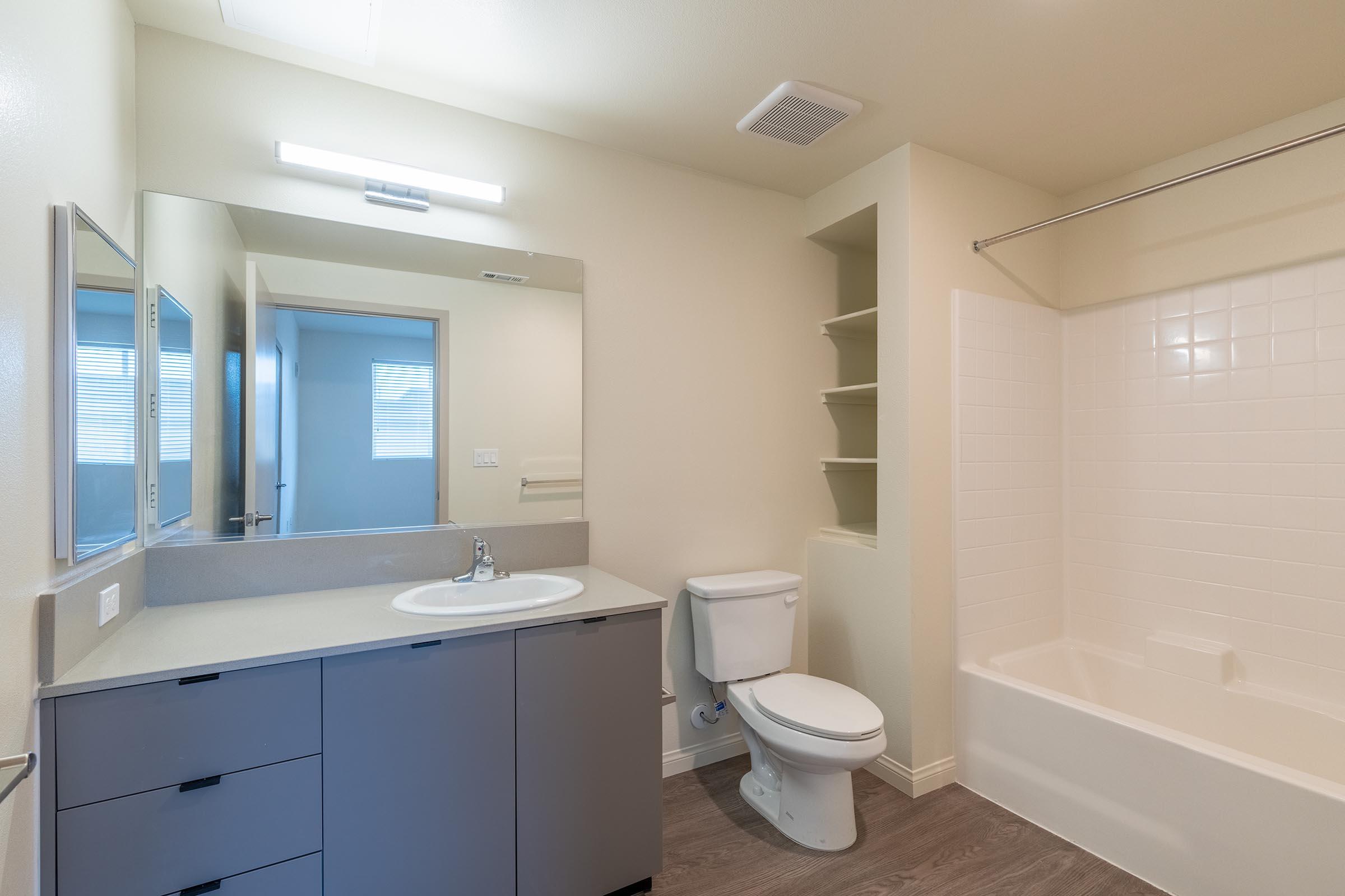 A modern bathroom featuring a white bathtub and shower, a light gray vanity with a sink, and a toilet. There is a large mirror above the sink, and natural light coming through a nearby window. The walls are a light cream color, and there is shelving next to the bathtub. The flooring is a wood-like texture.