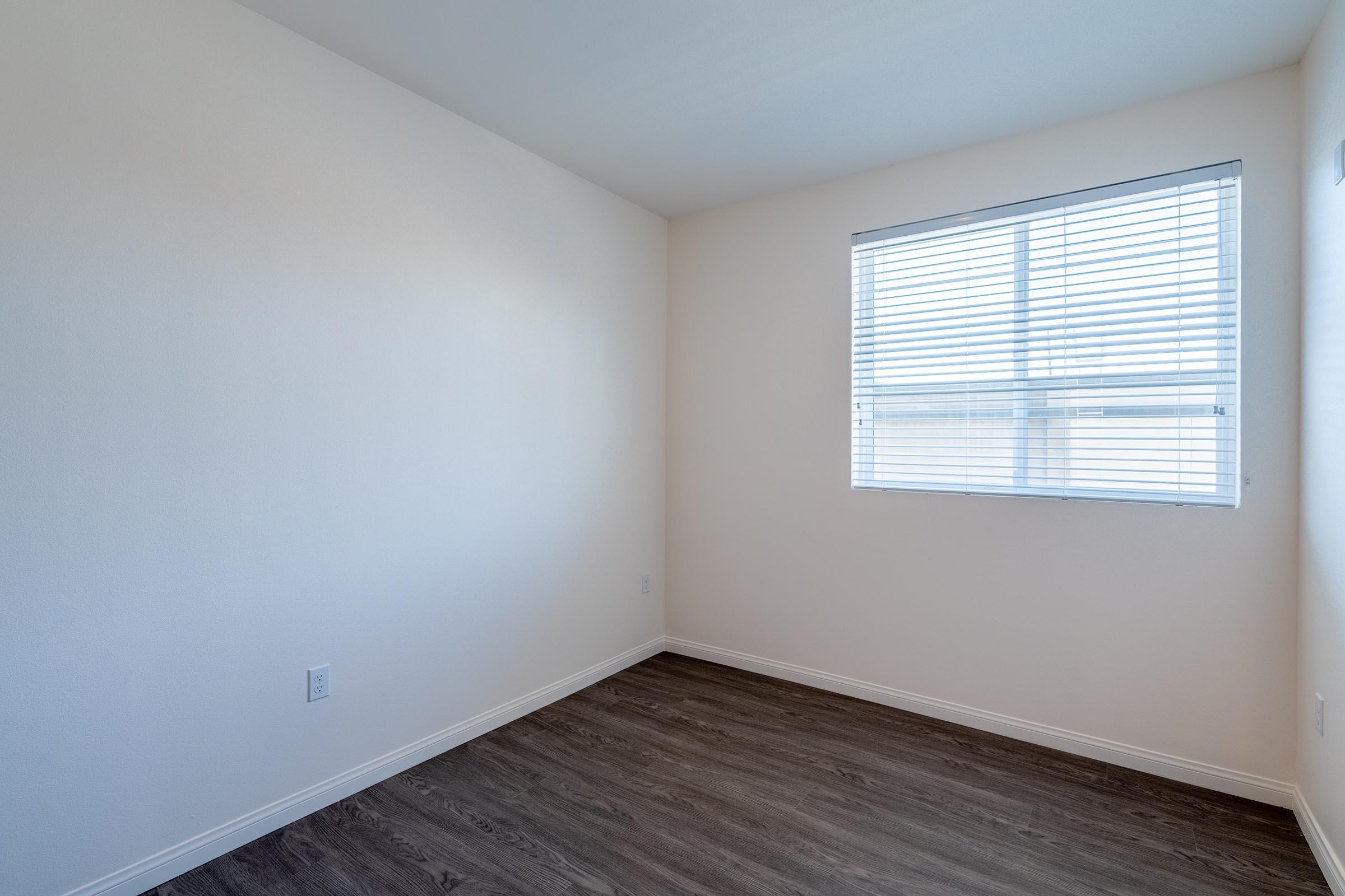 A small, empty room with light-colored walls and a wooden floor. There is a window with white blinds allowing natural light to enter. The room is sparsely furnished, creating an open and airy feel.