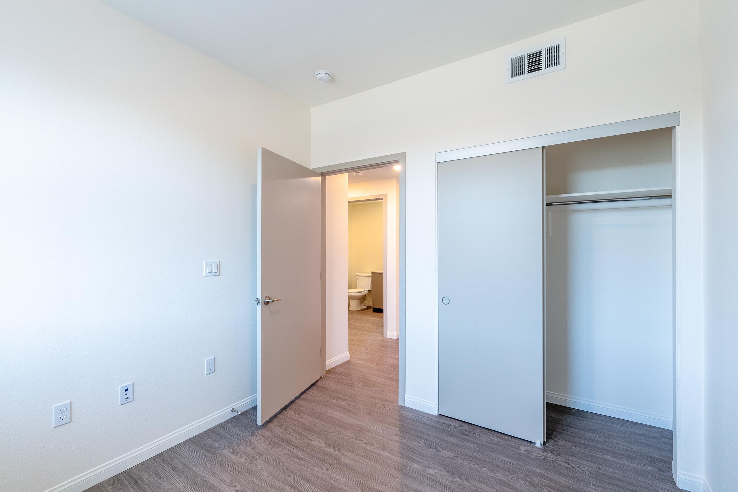A clean, empty room with light-colored walls and gray laminate flooring. A slightly open door on the left leads to a bathroom, while a closet with sliding doors is visible on the right. The overall atmosphere is bright and spacious, making it suitable for various interior designs.
