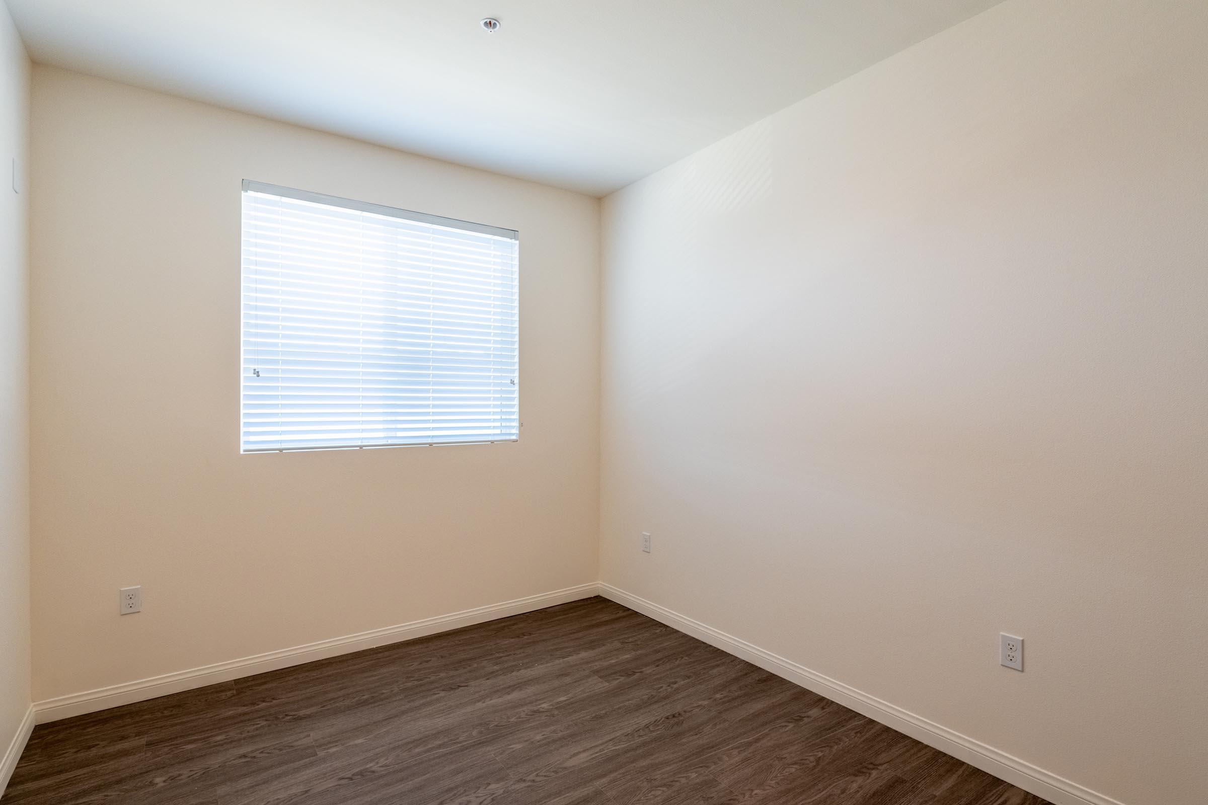 A blank room with light-colored walls and a window covered by white blinds. The floor is hardwood, and there are no furnishings or decorations visible, creating an open and minimalist atmosphere.