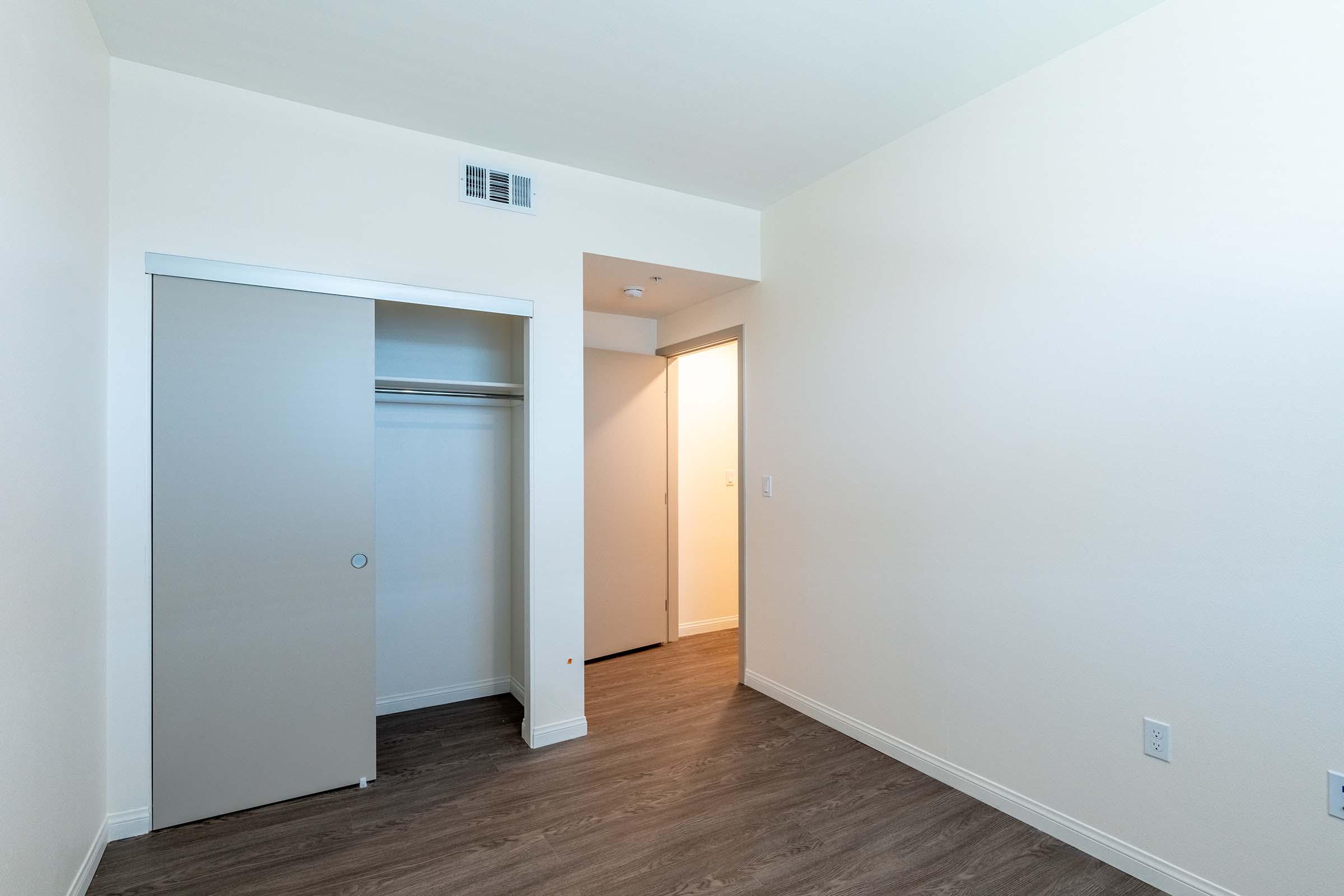 A small, empty room with light-colored walls and a wood-like floor. It features a sliding closet door on the left, a doorway leading to another room on the right, and a light source from the adjacent area. The overall space appears clean and unfurnished.