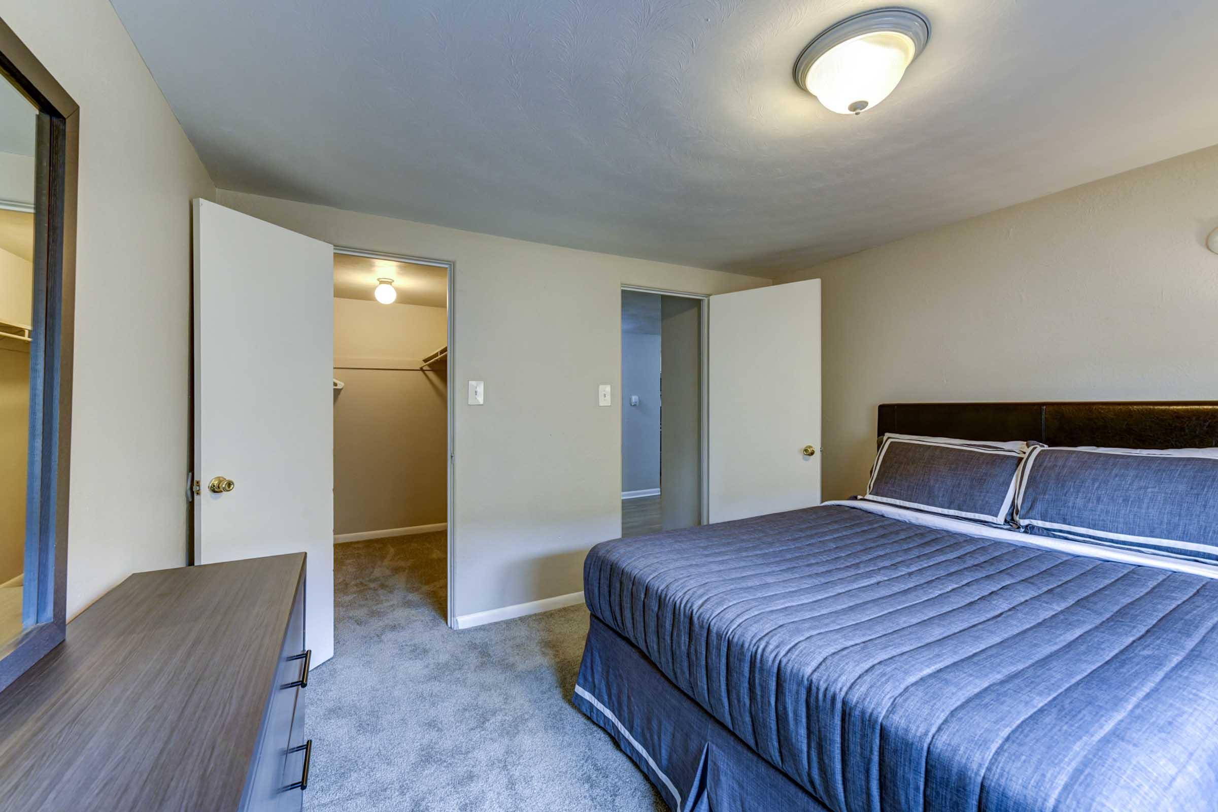  A well-lit bedroom featuring a grey upholstered bed with a neatly made blue bedspread. 