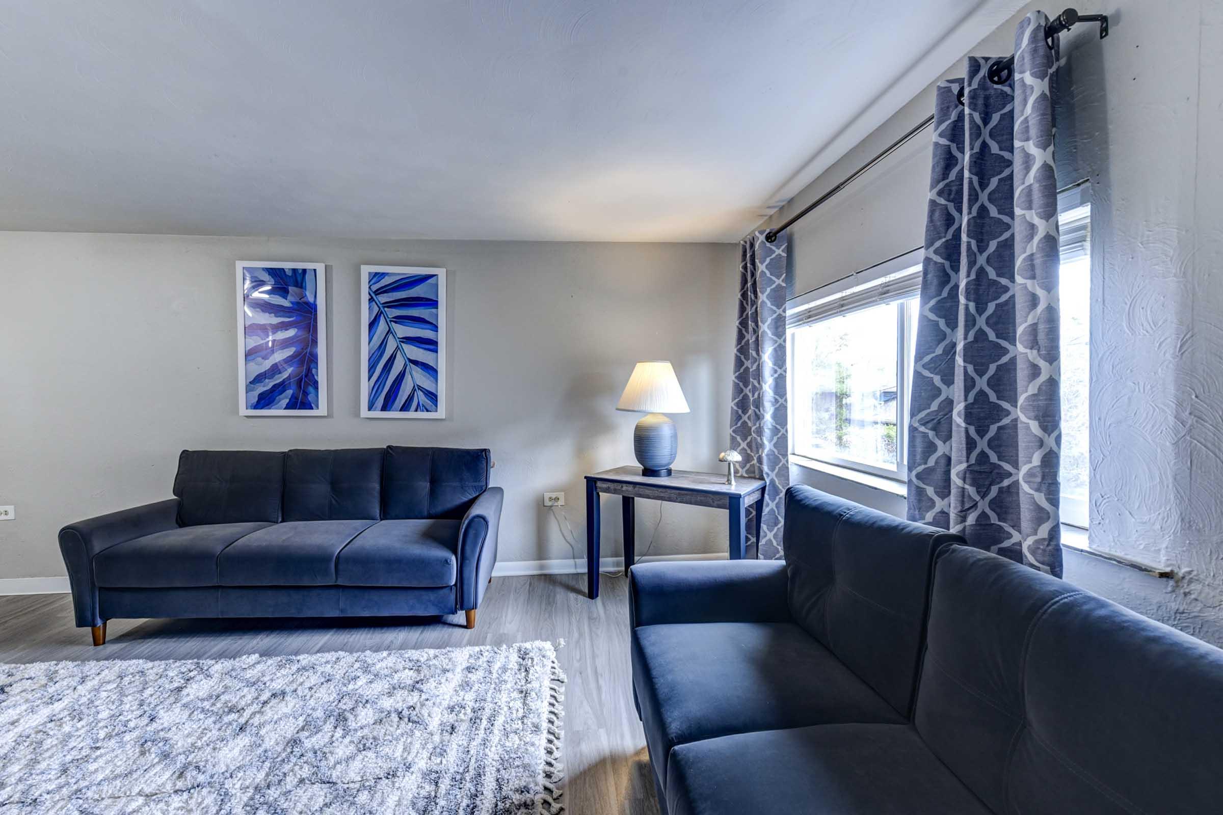 A modern living room featuring two dark blue sofas, a wooden side table with a lamp, and two framed artworks with blue designs on the walls. 