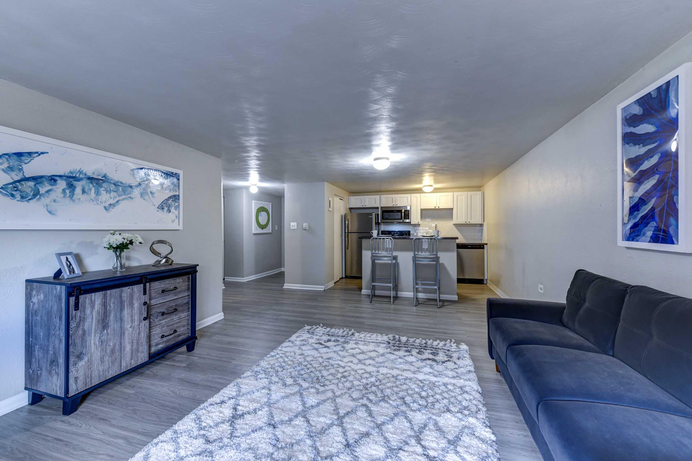 Spacious living area featuring a modern sofa, a decorative rug, and a sideboard. In the background, the open kitchen is visible, equipped with stainless steel appliances and white cabinetry. 