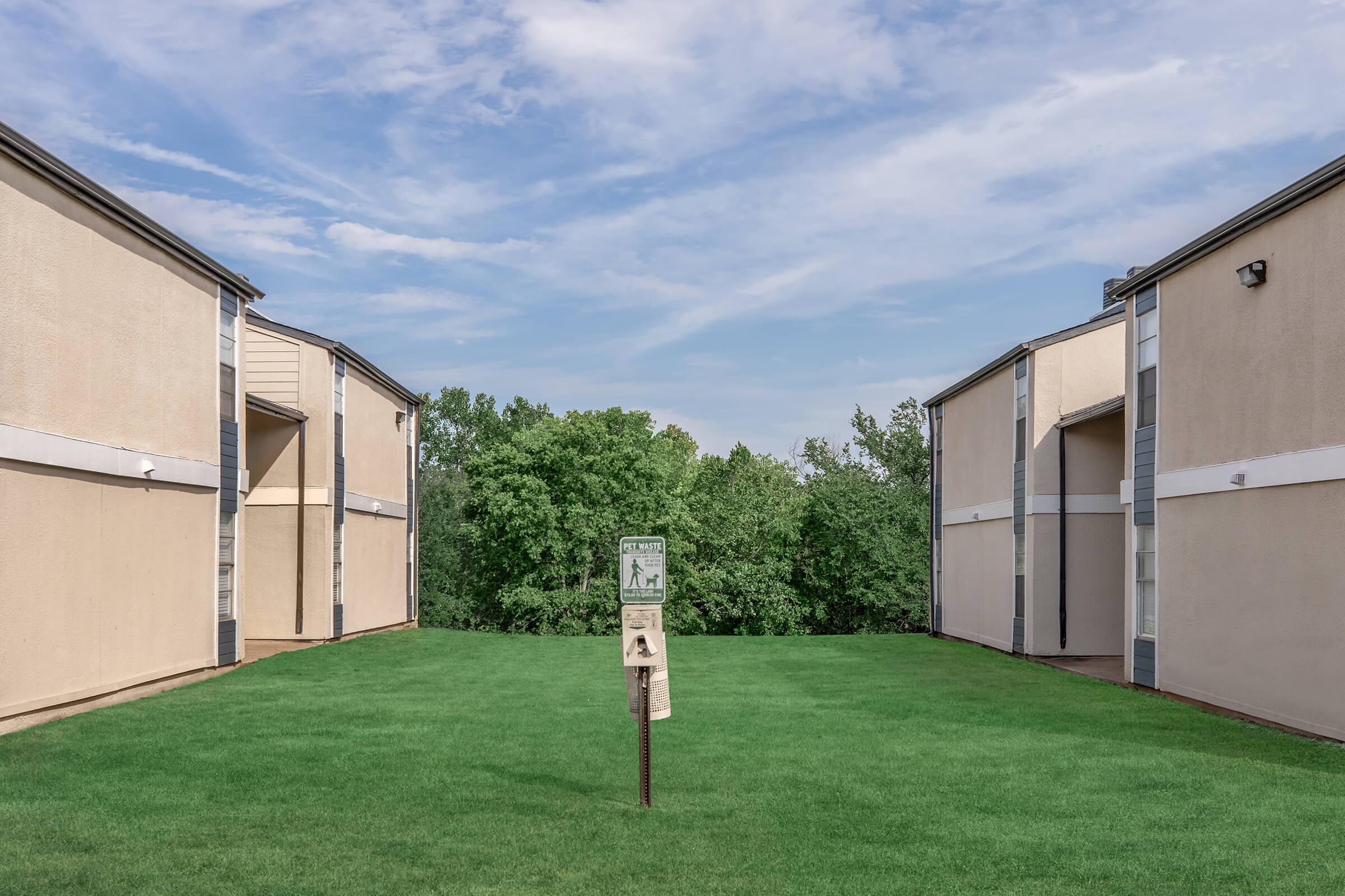A view between two apartment buildings, showcasing a well-maintained green lawn. In the center, a sign stands on a post, indicating itโs a pet-friendly area. The background features trees and a clear blue sky with some clouds. The overall atmosphere is calm and inviting.