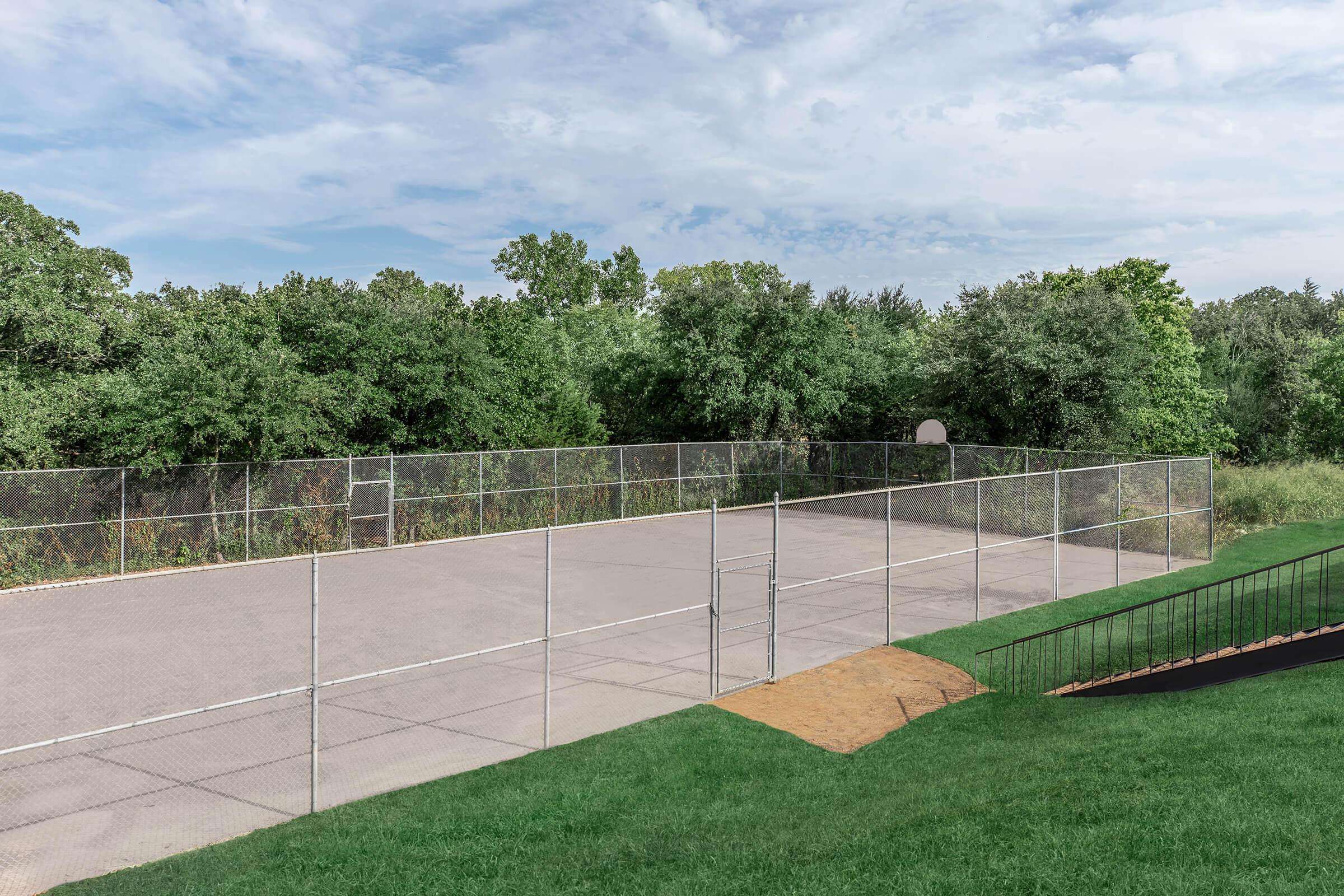 Empty outdoor basketball court enclosed by a wire fence, surrounded by lush greenery and trees under a partly cloudy sky. The court features a single basketball hoop and a well-maintained grassy area nearby.