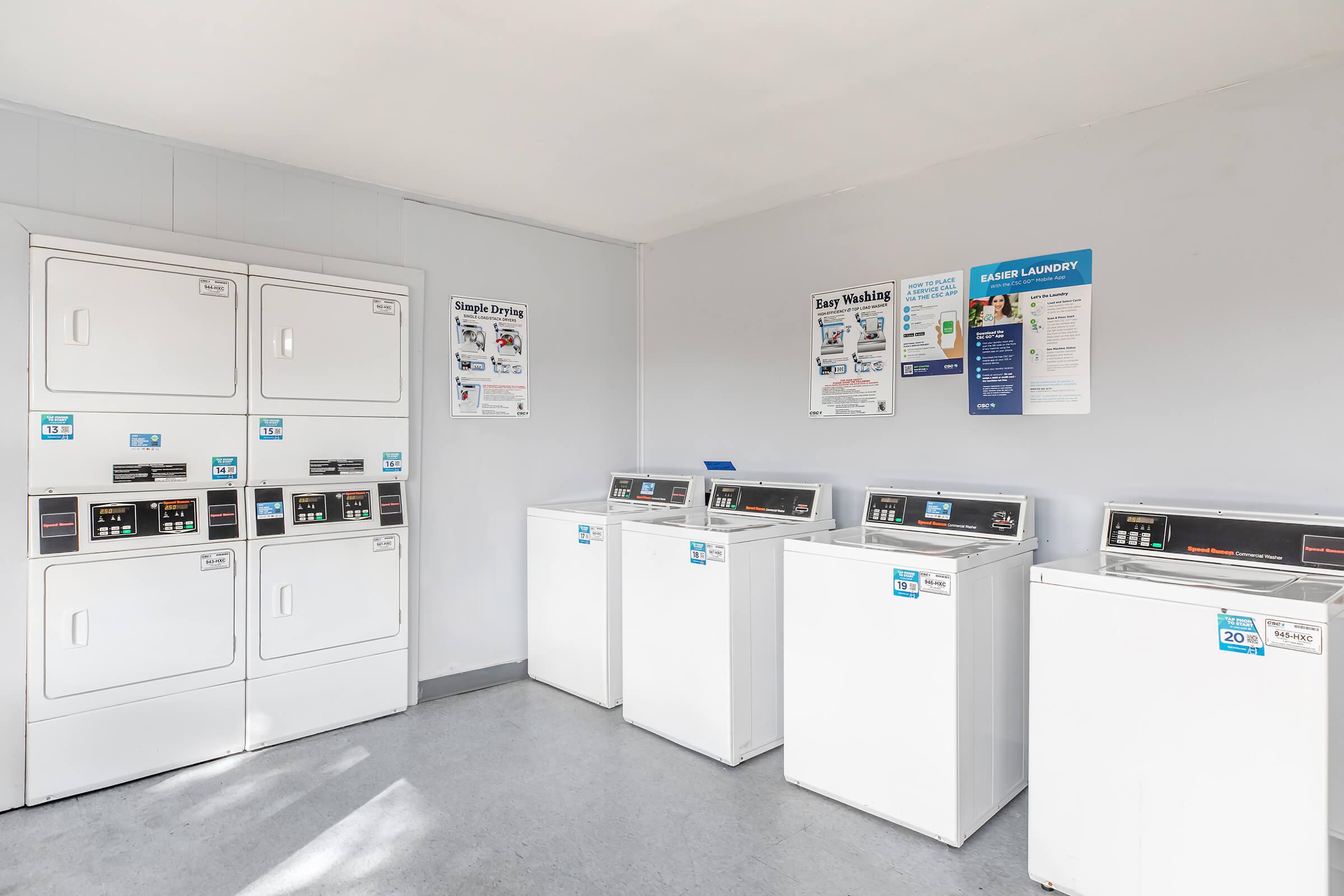 Interior of a laundromat featuring several white washing machines and dryers. There's a wall with informational posters on laundry services and instructions. The space is bright and clean, with gray flooring and a minimalist design, providing a functional area for laundry tasks.