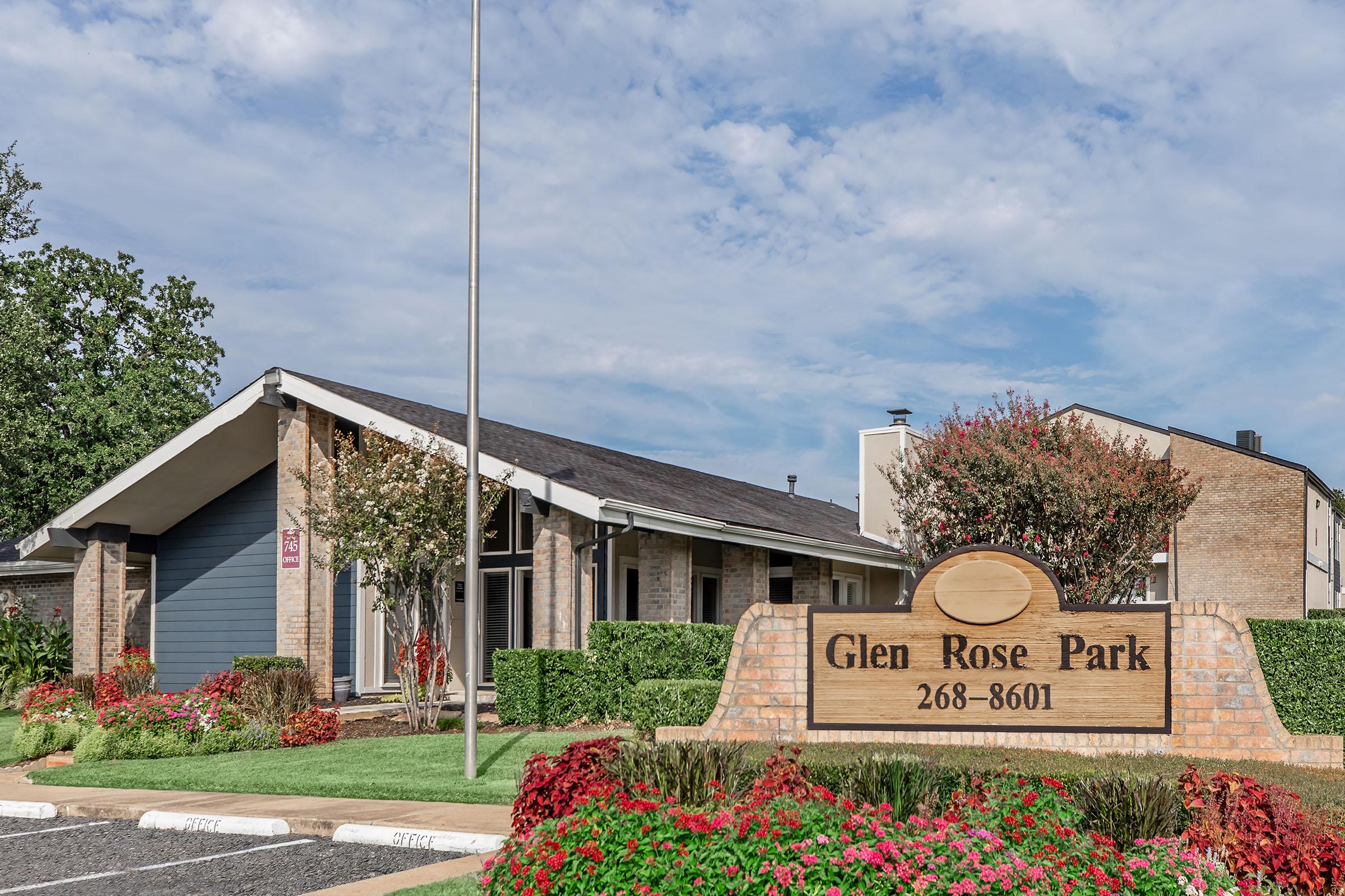 Sign for Glen Rose Park with the address 268-8601, surrounded by colorful flower beds and greenery. The building features a modern design with large windows and a sloping roof, set against a sky with light clouds. A flagpole stands in front of the building.