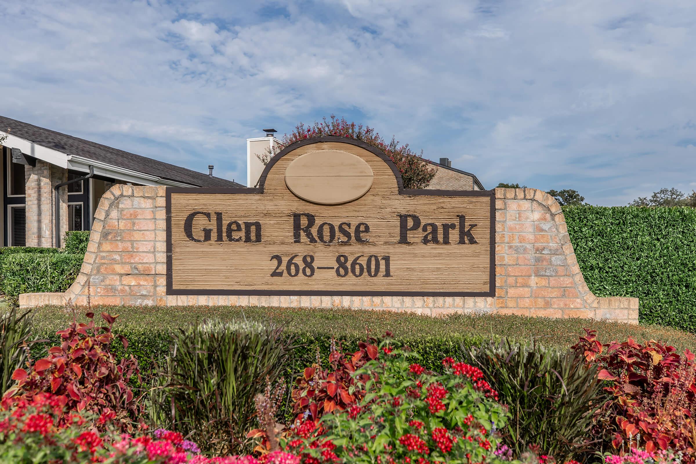 Sign for Glen Rose Park, featuring the park's name and phone number (268-8601), surrounded by colorful flowers and green landscaping. The background includes a building and a clear blue sky.