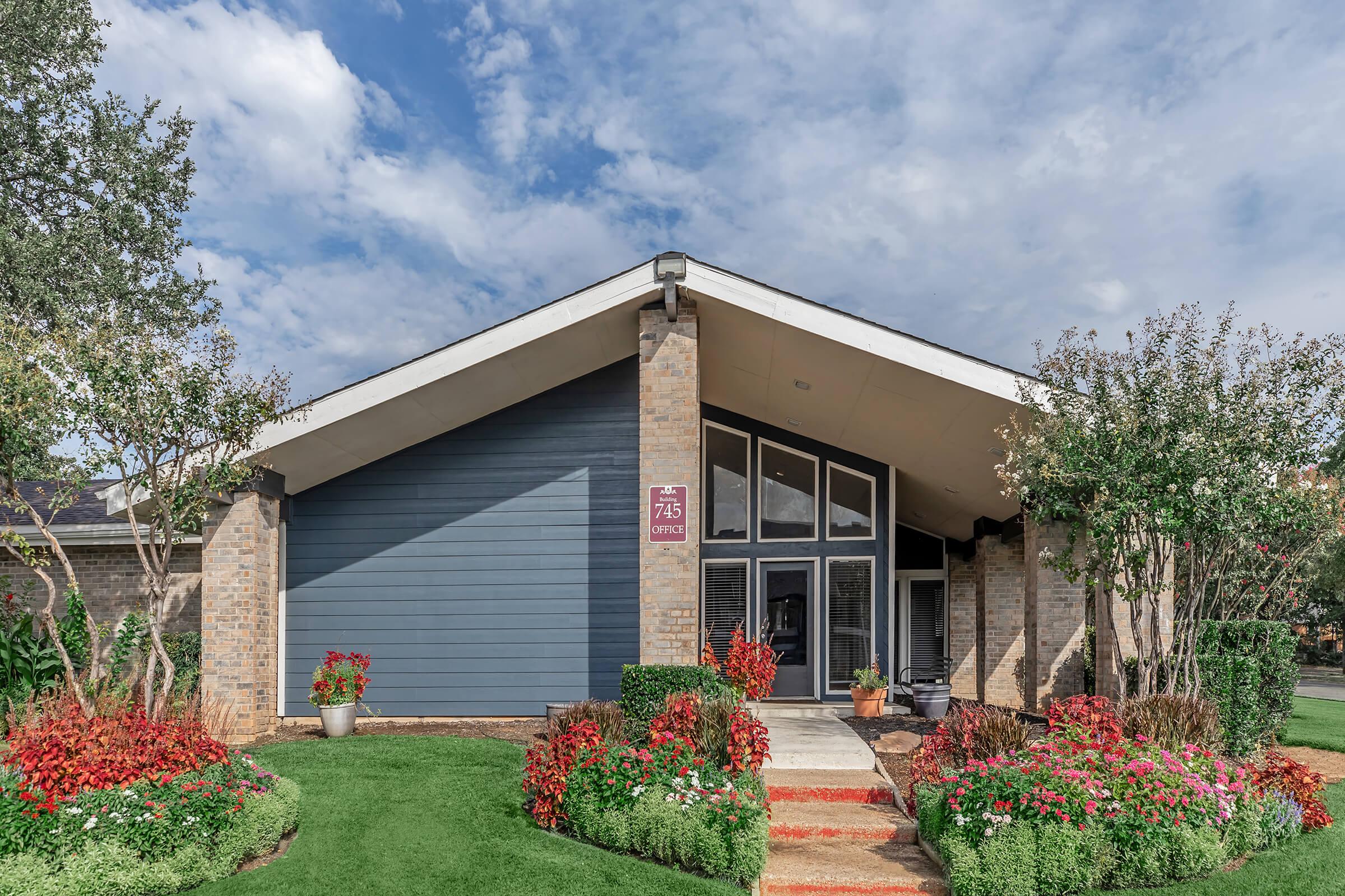 A modern building featuring a peaked roof with large windows, surrounded by colorful flower beds and greenery. The faรงade is a mix of brick and blue siding, and a sign indicates the building number. The sky is partly cloudy, adding to the bright atmosphere of the setting.