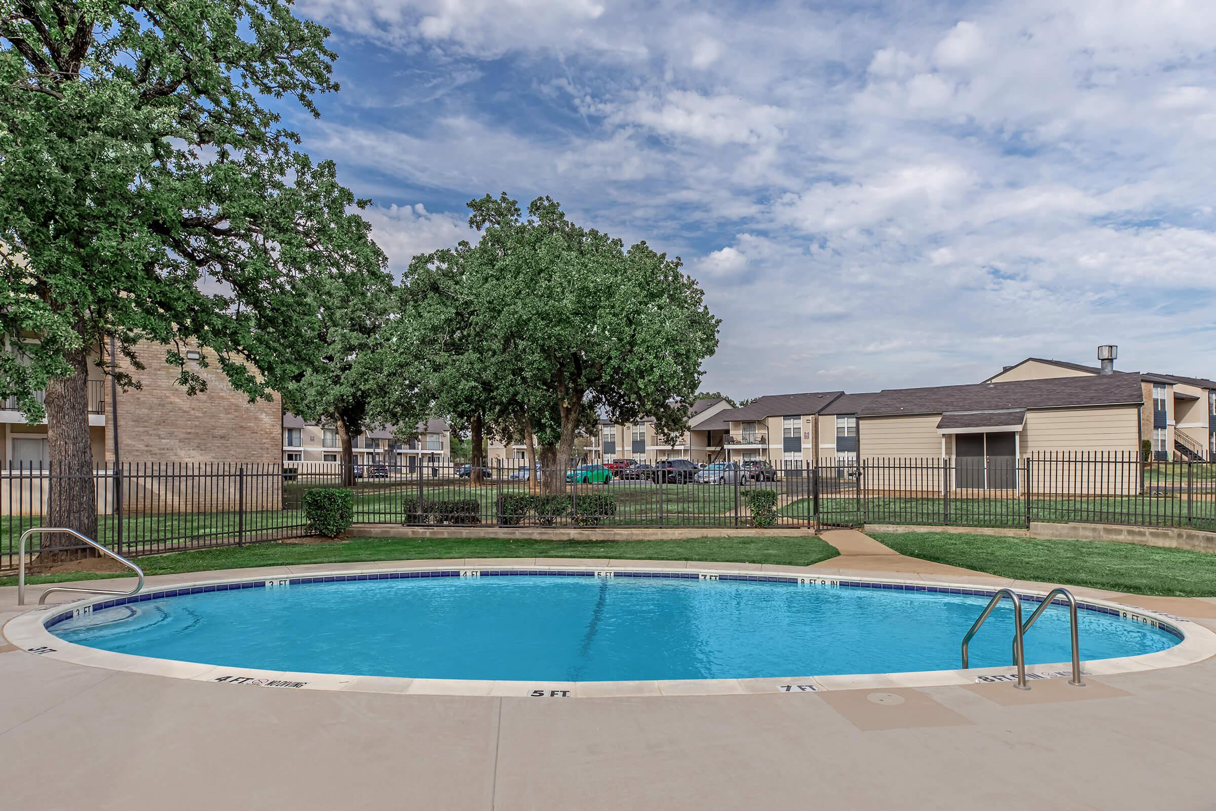 A well-maintained swimming pool in an apartment complex, surrounded by lush green trees and grassy areas. The sky is partly cloudy, adding a serene atmosphere to the scene. Several apartment buildings are visible in the background, indicating a residential setting.
