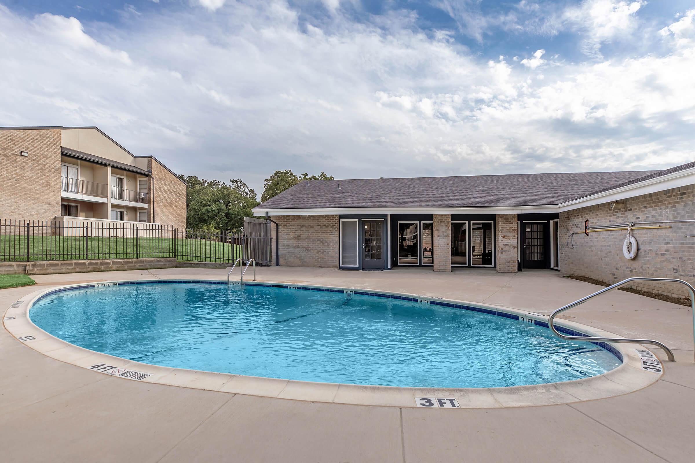 A rectangular swimming pool surrounded by a concrete deck, featuring steps for entry. In the background, a brick building with multiple floors and balconies is visible, along with a grassy area and cloudy sky. A lifebuoy hangs on the wall near the pool, and the depth markings are visible on the edge of the pool.